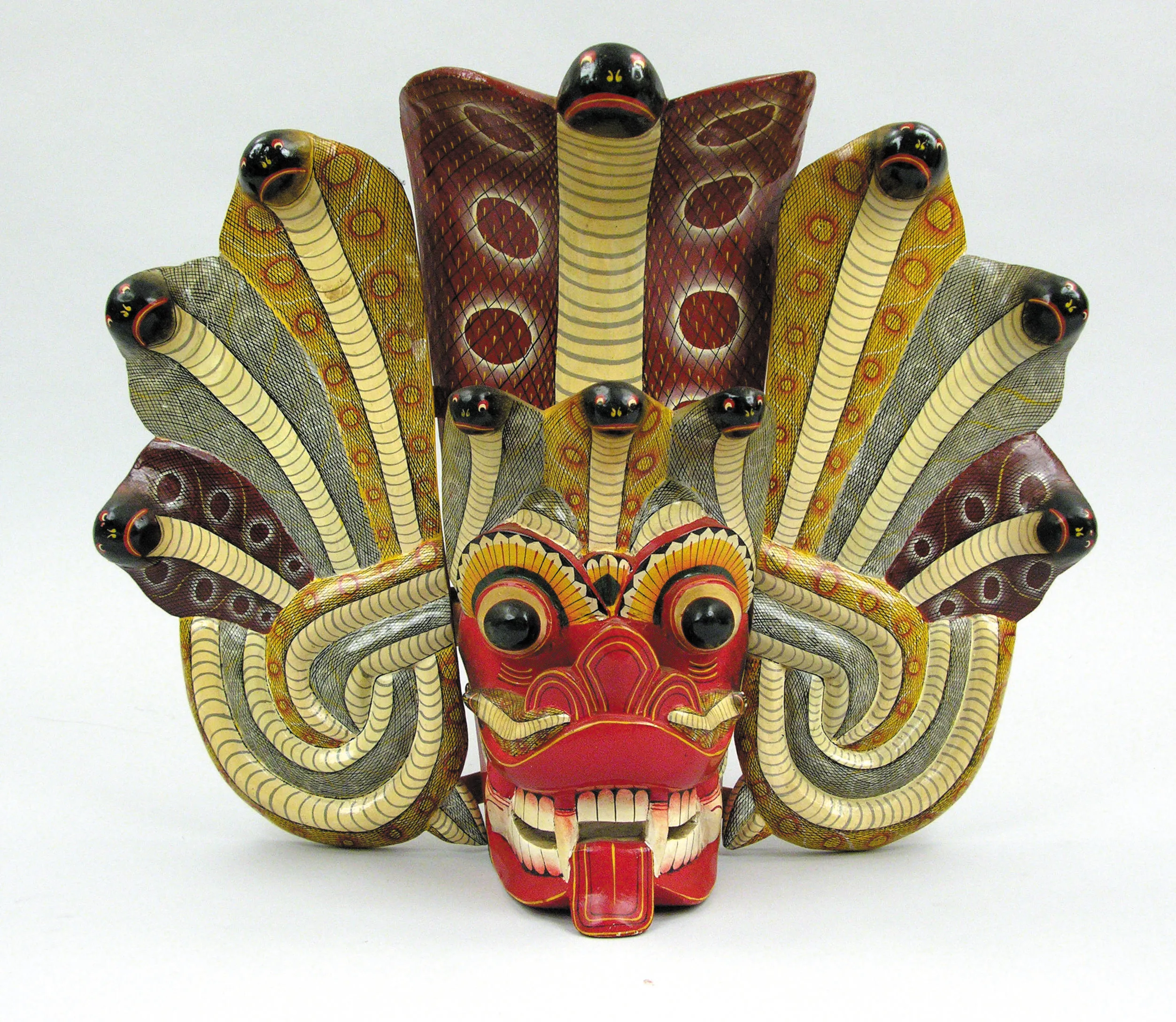 Una máscara de danza tradicional de Sri Lanka con cara roja, ojos negros y lengua protuberante, rodeada de múltiples figuras serpentinas con patrones amarillos y verdes.