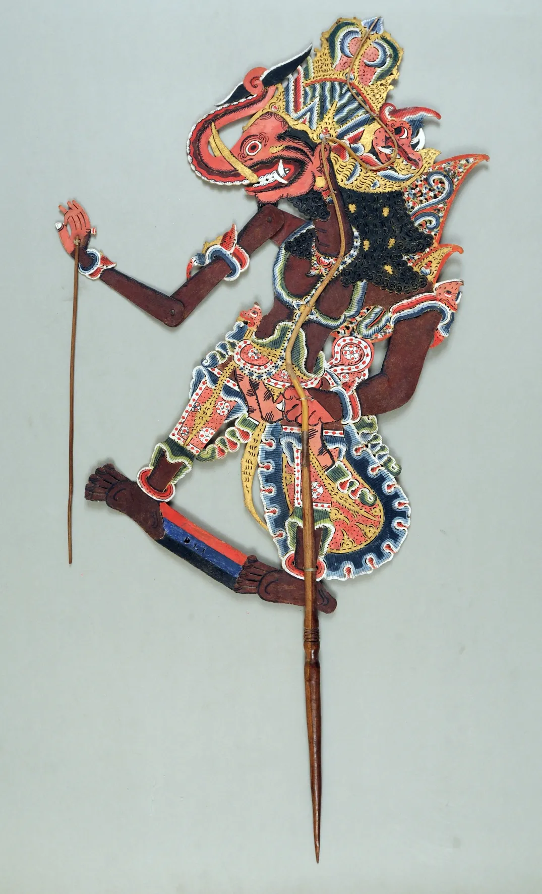 Una detallada marioneta de sombras Wayang Kulit con cara roja, tocado ornamentado y patrones coloridos, sosteniendo una vara contra un fondo liso