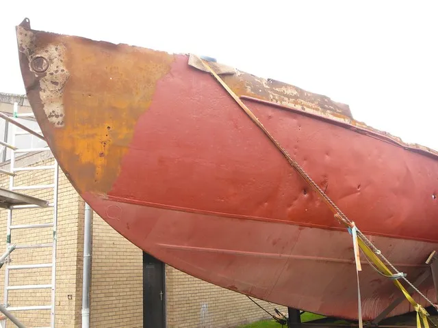 Casco de color rojo y óxido de un velero sobre soportes con andamios visibles al fondo