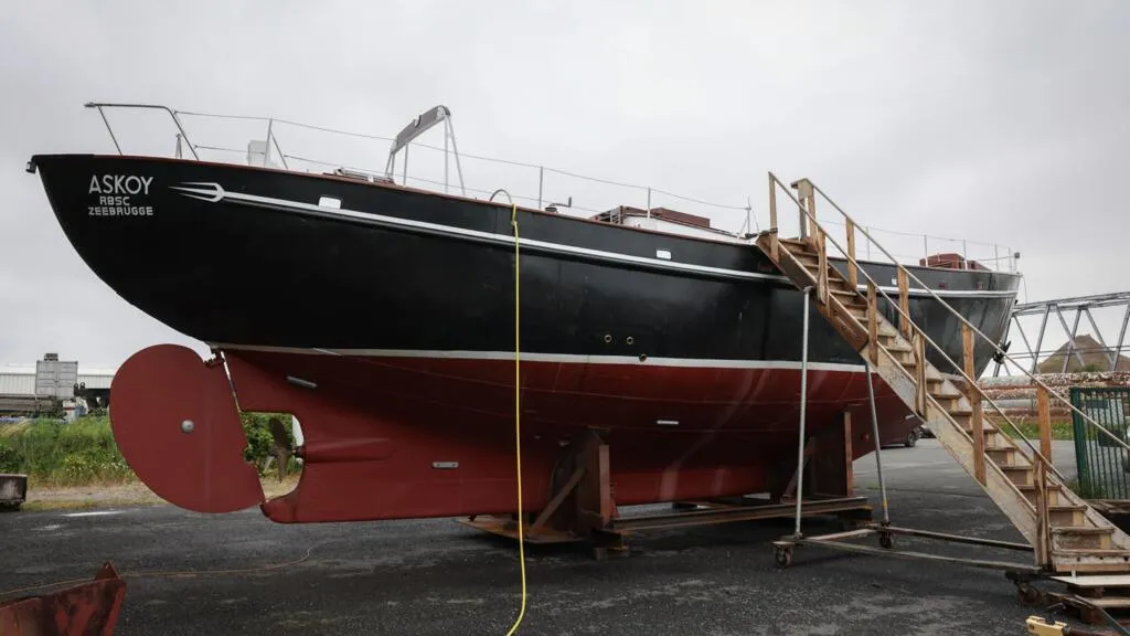 El velero negro y rojo Askoy II reposando sobre soportes en un astillero, con una escalera de madera que conduce a su cubierta