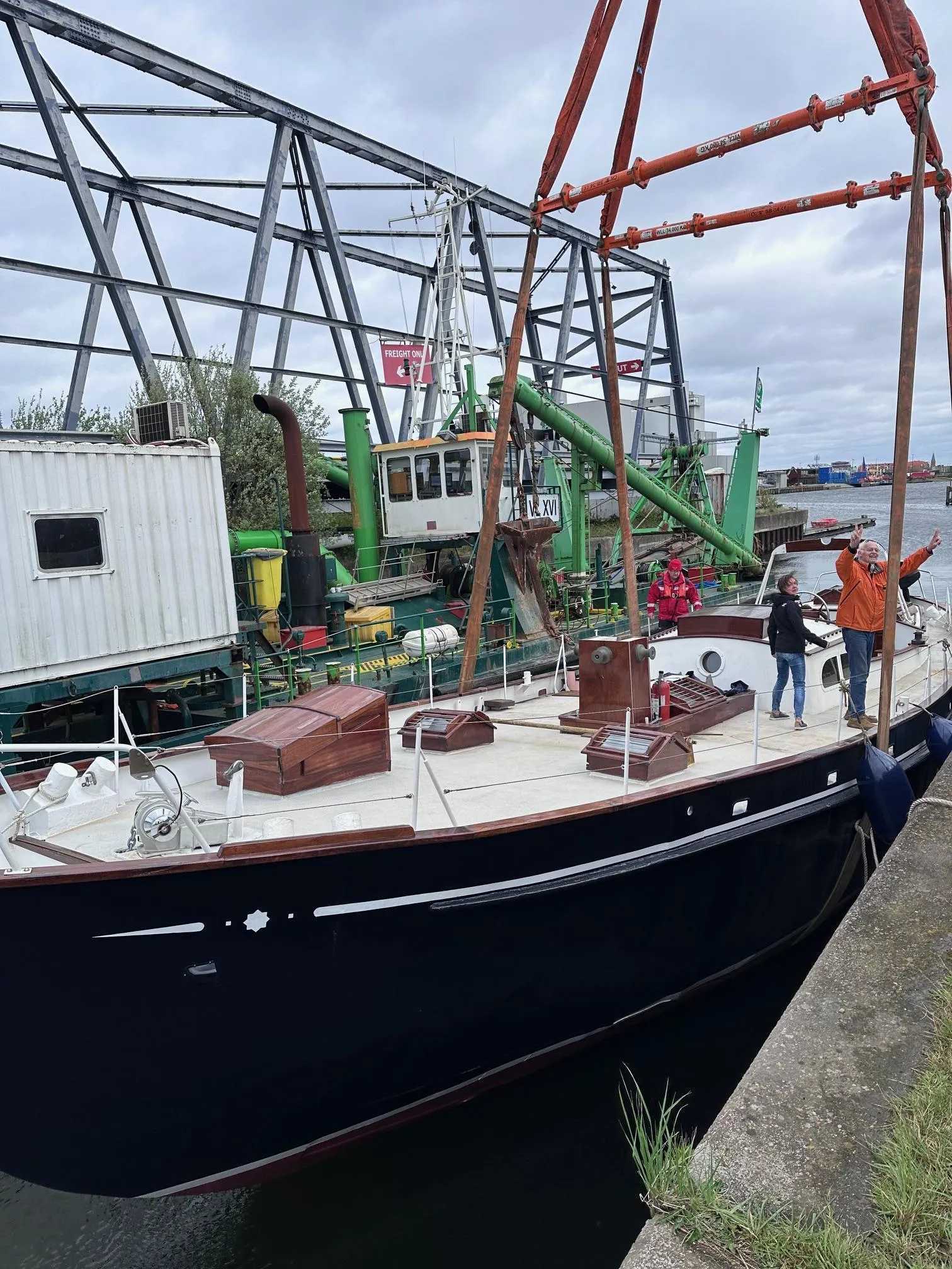 Velero de casco oscuro atracado en un puerto con visitantes de pie en la cubierta blanca, con una gran estructura de puente metálico y equipo industrial al fondo