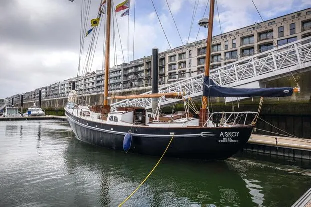 El velero de dos mástiles y casco oscuro ASKOY atracado en un puerto deportivo con modernos edificios de apartamentos y un puente peatonal blanco al fondo.