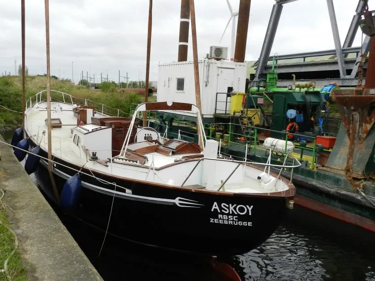 Velero de casco negro llamado ASKOY con la inscripción 1952 ZEEBRUGGE en la proa, atracado en un puerto con mástiles, defensas azules y equipo industrial al fondo