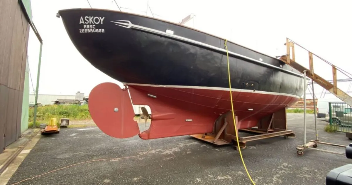Casco de un velero negro y rojo llamado ASKOY descansando sobre una cuna en un astillero, mostrando el timón y la hélice.