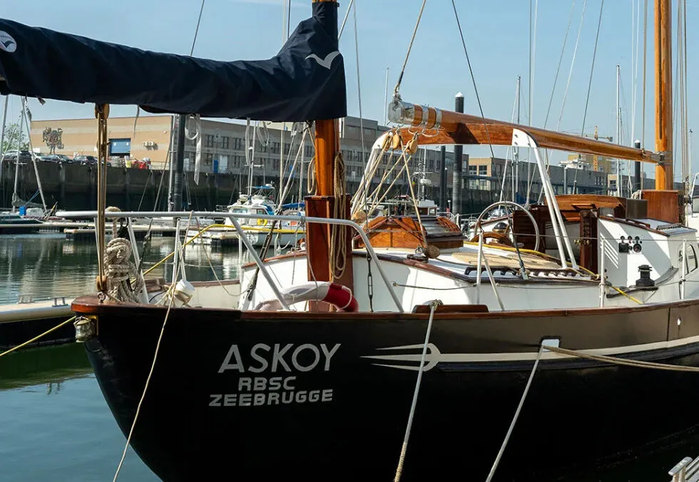 Casco de un barco de vela negro con el texto en blanco ASKOY ABSC ZEEBRUGGE, atracado en un puerto deportivo con mástil de madera y aparejos visibles