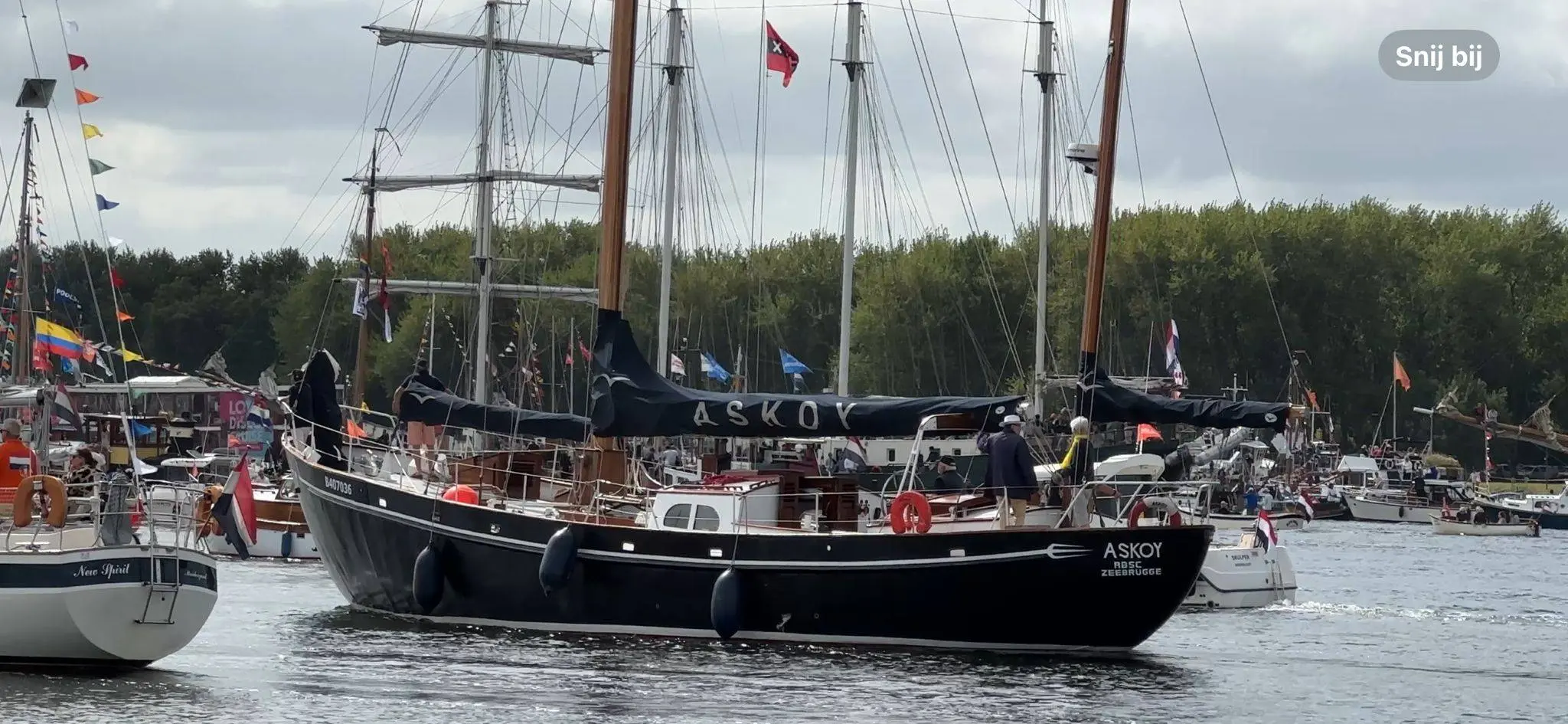 Velero negro de dos mástiles Askoy II en un puerto rodeado de otros barcos con una orilla arbolada al fondo