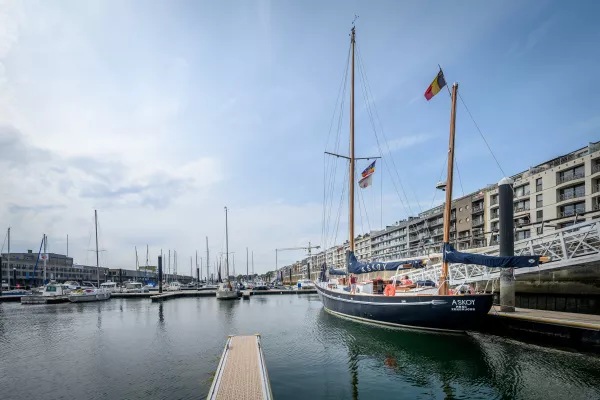 El velero Askoy II amarrado en un puerto deportivo, con su casco oscuro, sus altos mástiles de madera y sus banderas belgas visibles desde el otro lado del agua.