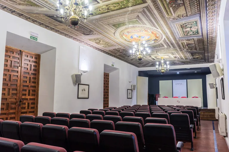 Auditorio con filas de sillas tapizadas oscuras frente a un escenario con una pantalla de proyección, bajo un techo ornamentado y pintado con lámparas de araña de cristal y puertas de madera con paneles a lo largo de las paredes