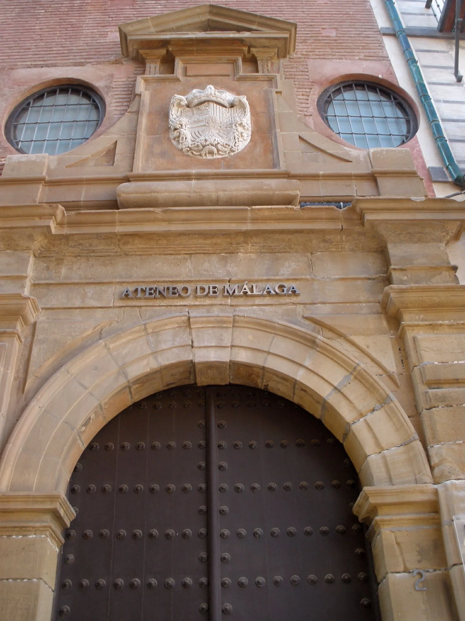 Kamienna fasada Ateneo de Málaga z łukowatymi drewnianymi drzwiami z metalowymi ćwiekami, rzeźbionym herbem i nazwą instytucji wyrytą nad wejściem