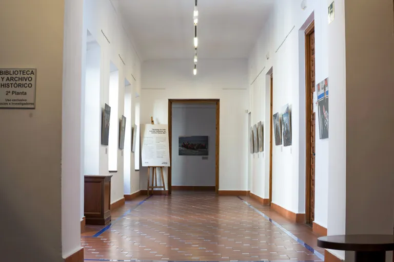 Un pasillo largo y blanco con obras de arte expuestas en las paredes, suelo de baldosas y un cartel que dice Biblioteca y Archivo Histórico en la pared izquierda.