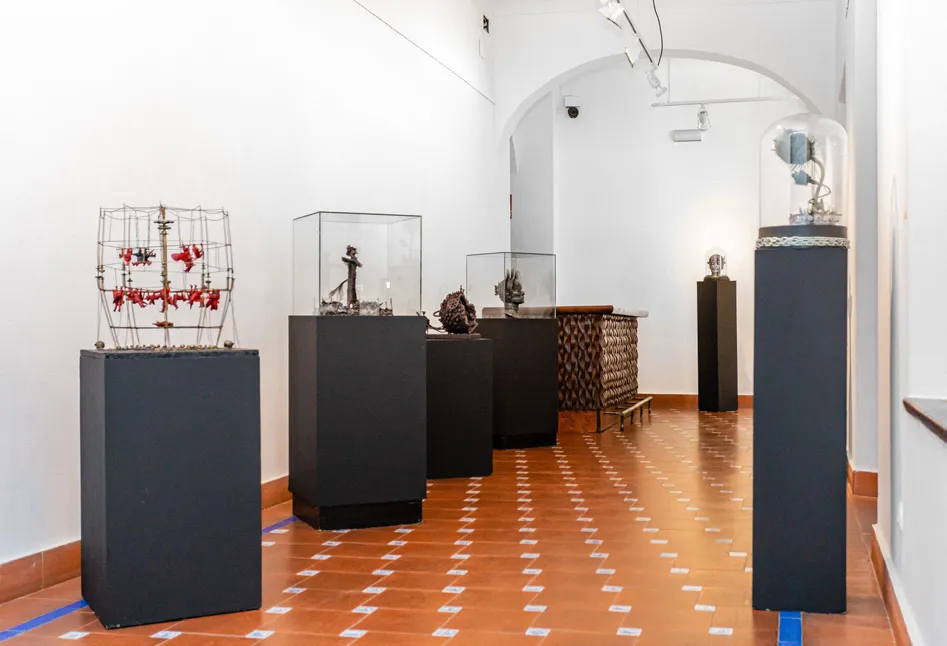 Sala de exposiciones interior con paredes blancas y suelo de baldosas de terracota, con varias esculturas sobre pedestales negros y en vitrinas de cristal.
