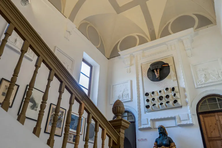 Interior de un edificio histórico que muestra una escalera de madera con balaustres, un techo abovedado con patrones geométricos, obras de arte enmarcadas en la pared y una instalación mural con un sombrero y elementos circulares sobre una escultura de un busto.