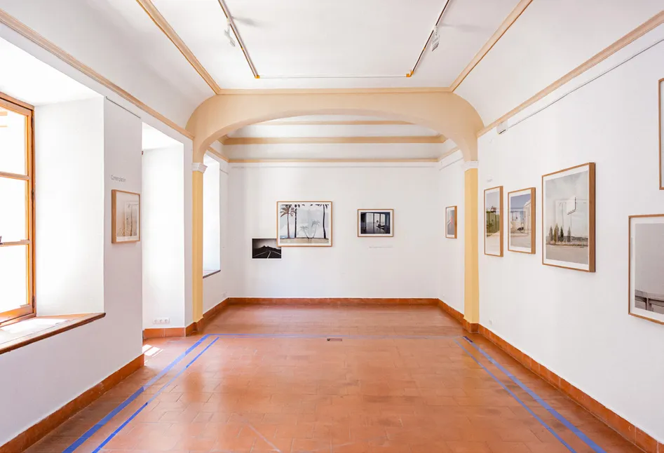 Una galería luminosa con paredes blancas, puertas arqueadas, suelo de terracota, iluminación de riel y obras de arte enmarcadas expuestas a lo largo de las paredes.