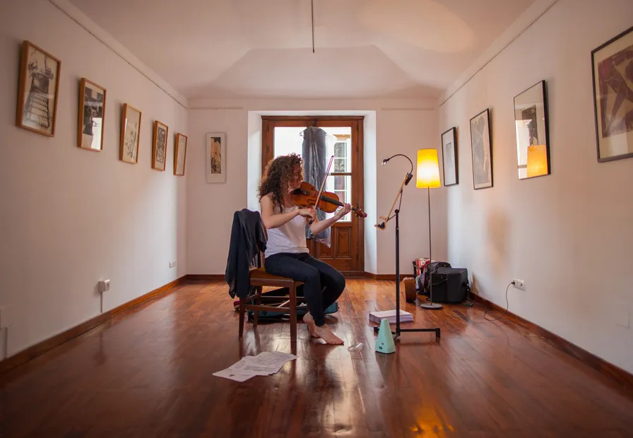 Una mujer tocando el violín, sentada en una larga galería con suelos de madera y obras de arte enmarcadas en las paredes.