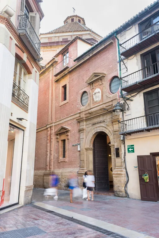 Fachada histórica de ladrillo rojo con una entrada de arco de piedra, dos ventanas circulares, un escudo de armas sobre la entrada y una cúpula de azulejos visible detrás del tejado, con gente caminando por la calle adoquinada frente al edificio.