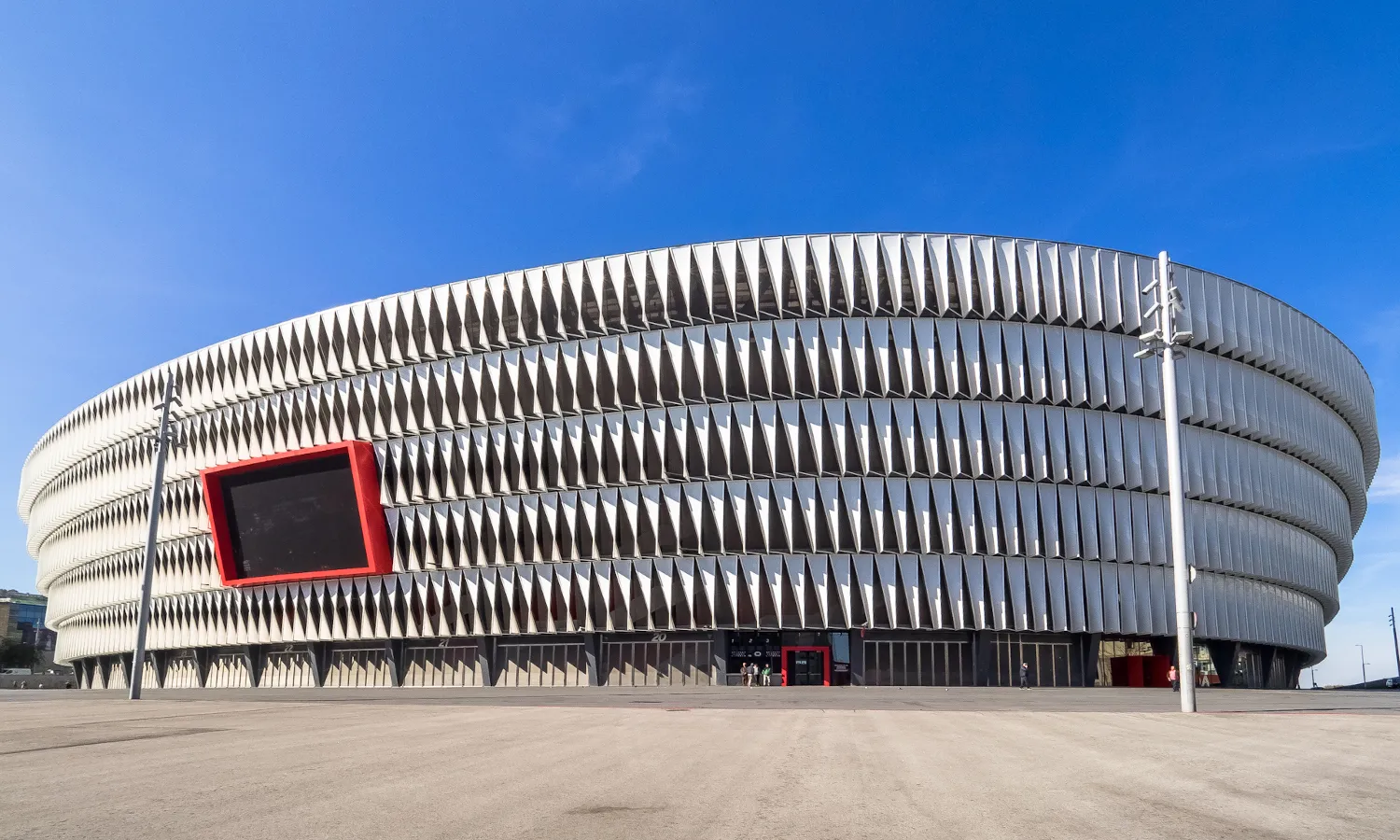 Totaalbeeld van de buitenkant van het moderne San Mamés-stadion met een gebogen, wit geribbelde gevel en een groot scherm met een rode omlijsting onder een helderblauwe lucht.