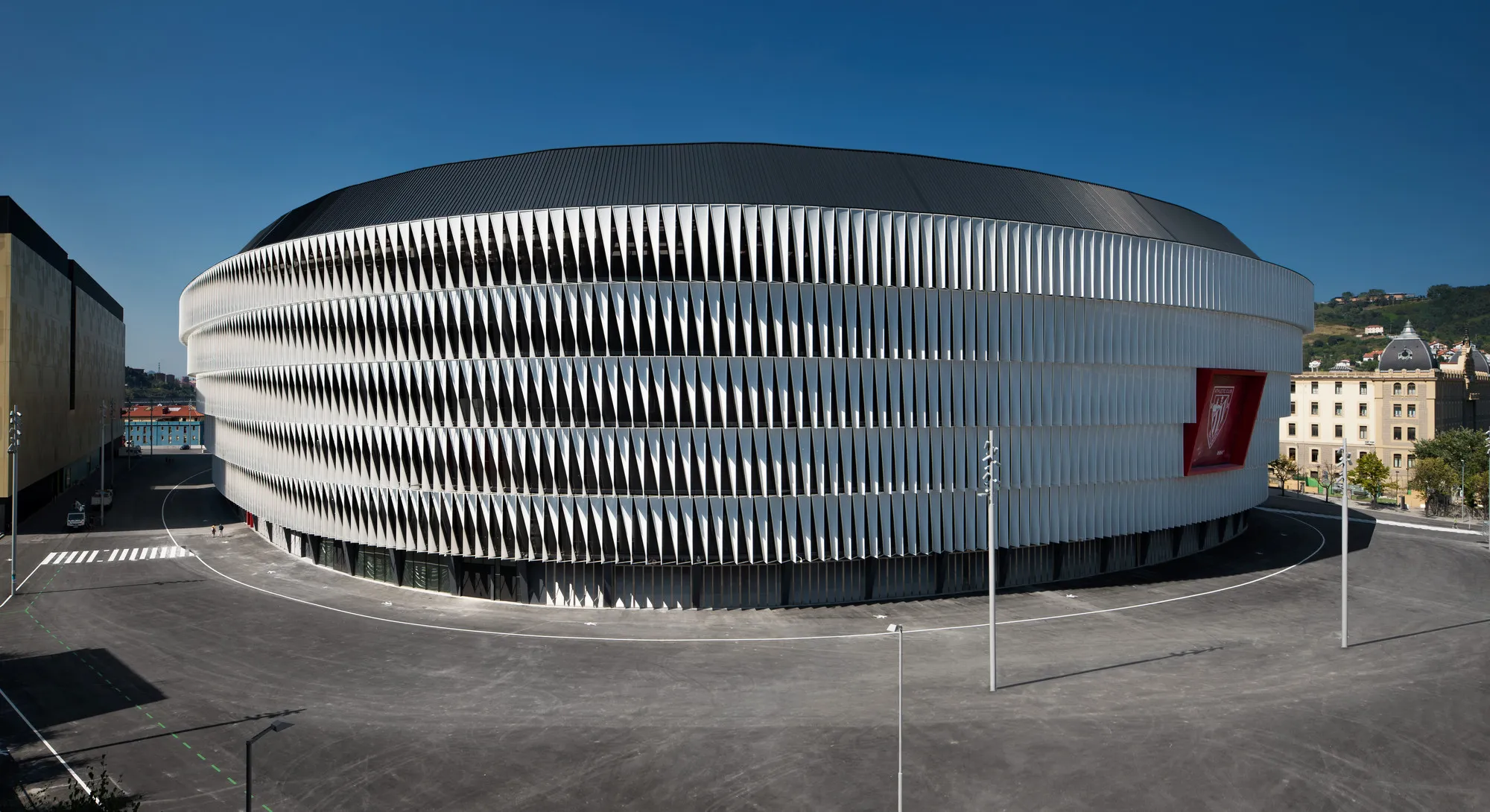 Breed exterieurbeeld van het San Mamés-stadion met de kenmerkende witte, gebogen gevel met verticale vinnen en het rode logo van Athletic Club