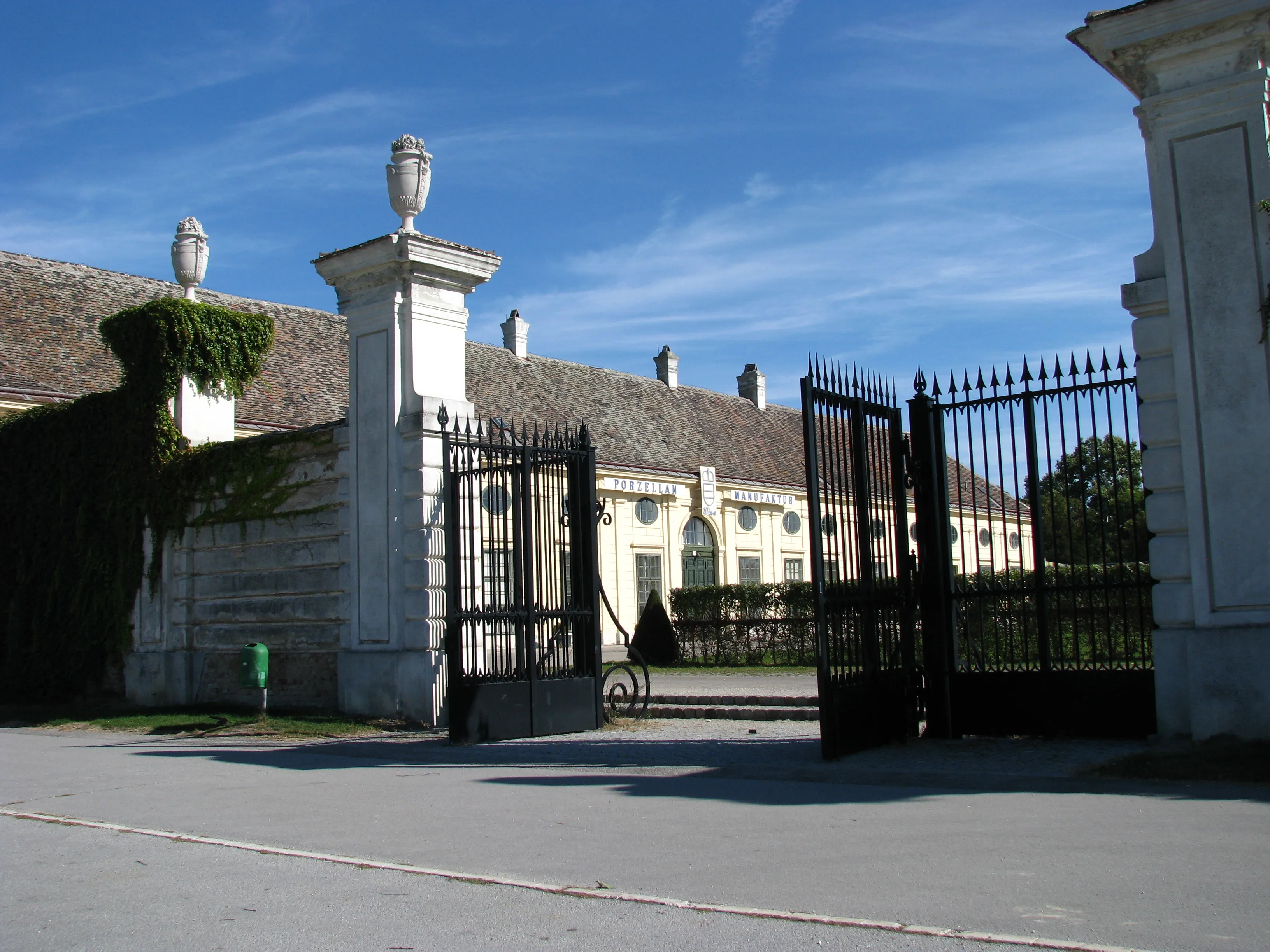 Puerta de piedra blanca con vallas de hierro negro y un edificio clásico con tejado de tejas y urnas decorativas