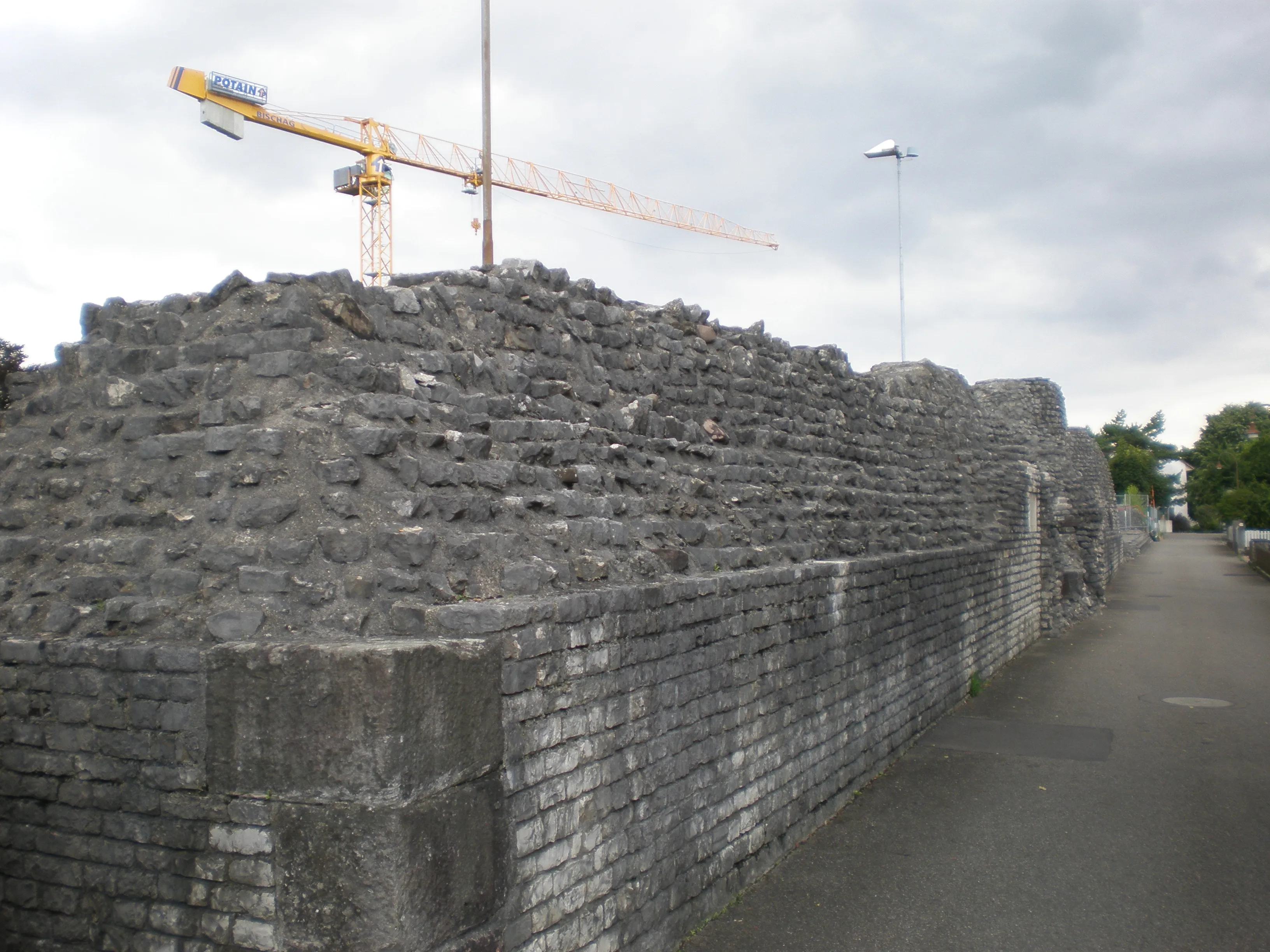 Ruïnes van een grote stenen muur op de archeologische vindplaats Augusta Raurica, met een bouwkraan erboven en een geplaveid pad ernaast.