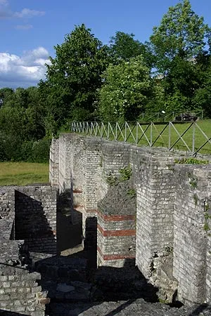 Oude Romeinse stenen ruïnes met gelaagde bakstenen muren en een houten hekwerk, omringd door groene bomen onder een blauwe lucht