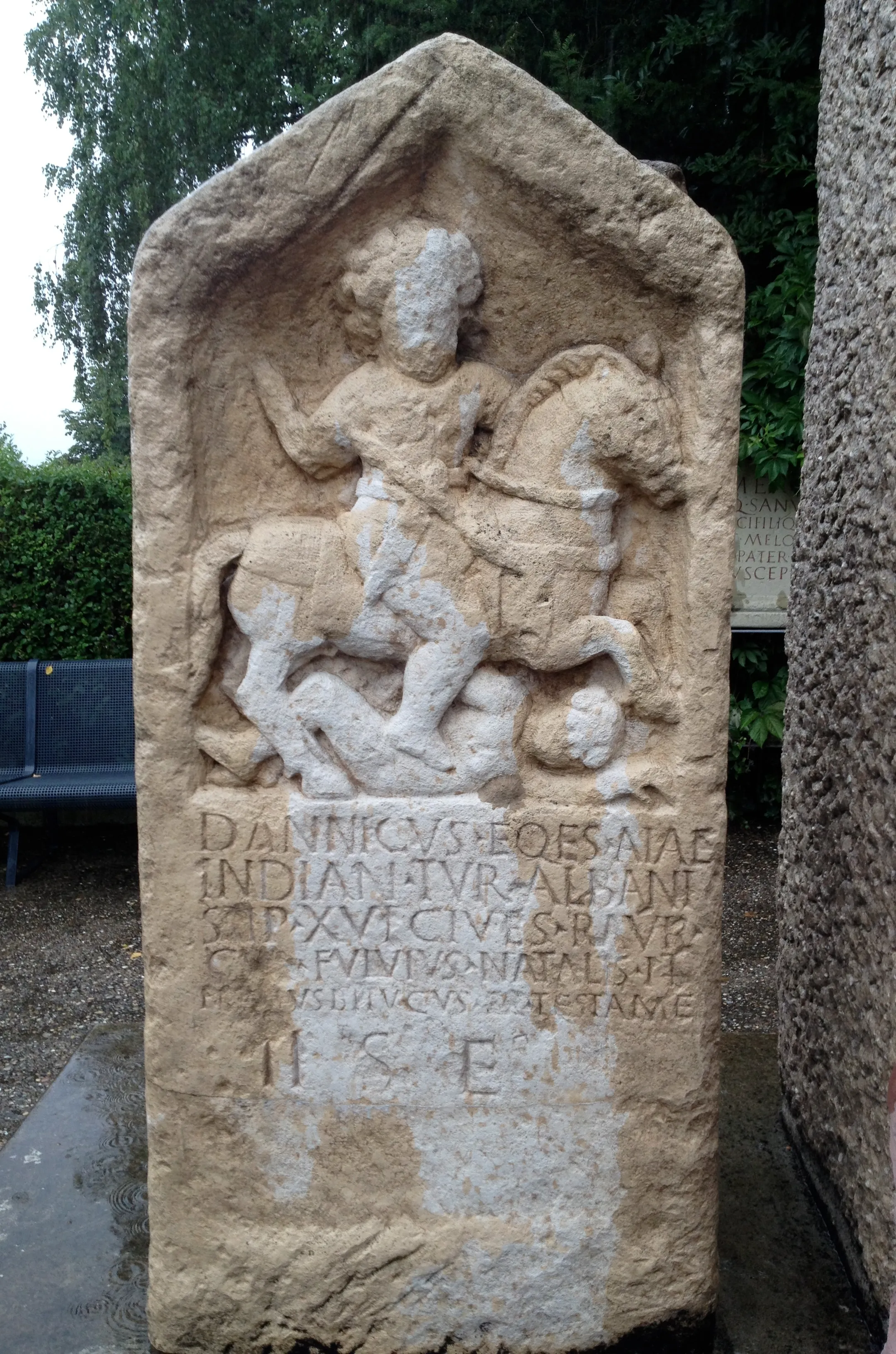 Romeinse stenen grafreliëf waarop een ruiter te paard over een gevallen figuur rijdt, met inscripties in het Latijn eronder, tentoongesteld in de openlucht in het lapidarium van Augusta Raurica.