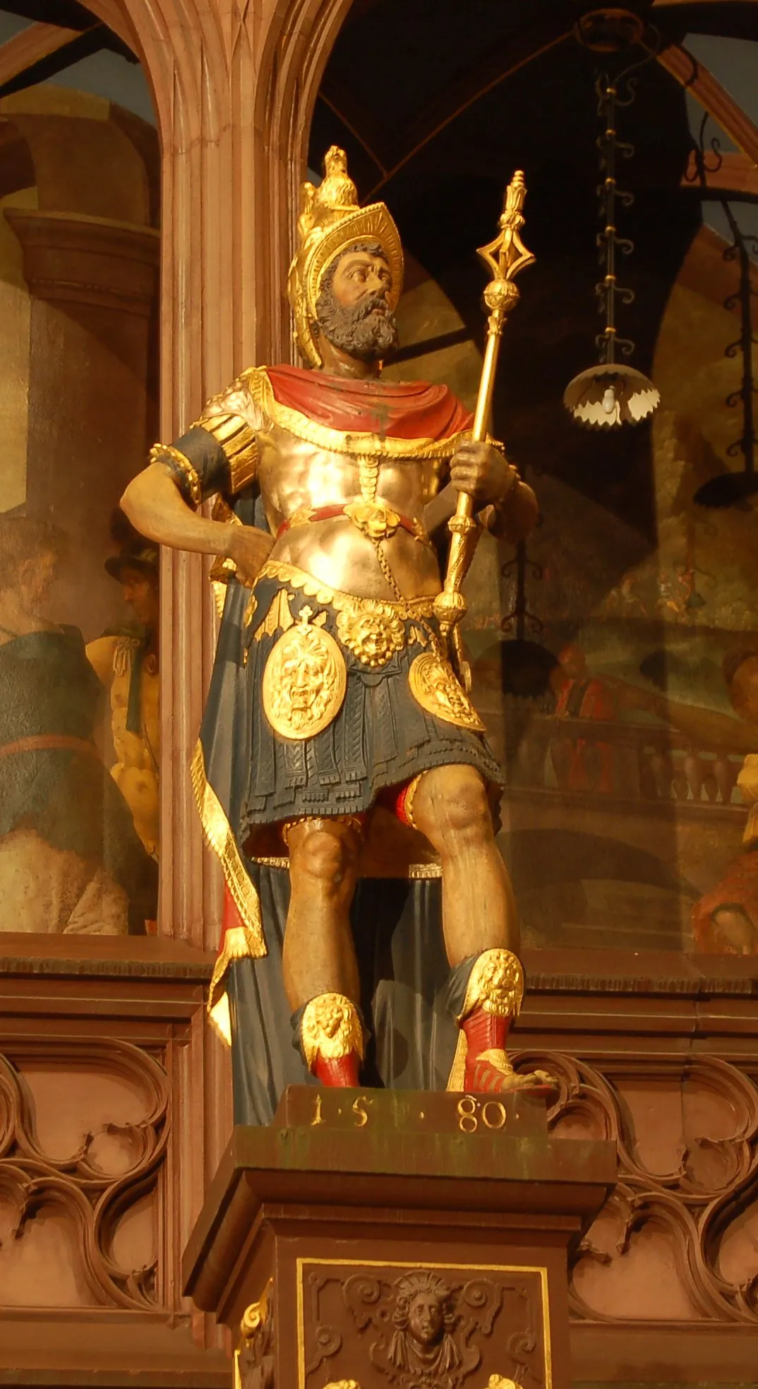 Vergulde standbeeld van een Romeinse soldaat in harnas met rode mantel, vasthoudend aan een gouden staf, staand op een voetstuk met het jaartal 1580.