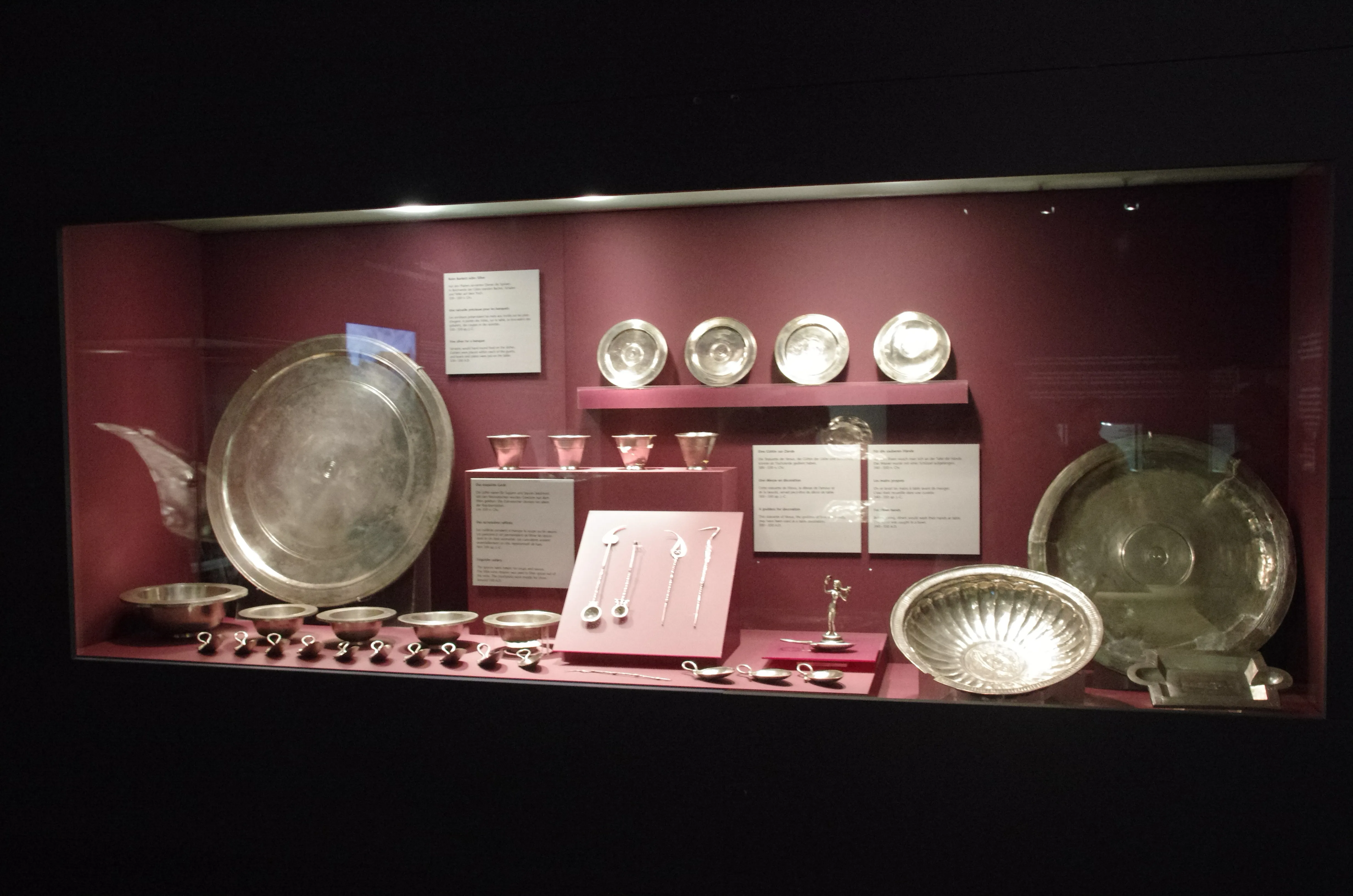 Museumvitrine met de Zilveren Schat van Augusta Raurica, met grote zilveren borden, schalen en bekers op planken tegen een bordeauxrode achtergrond.