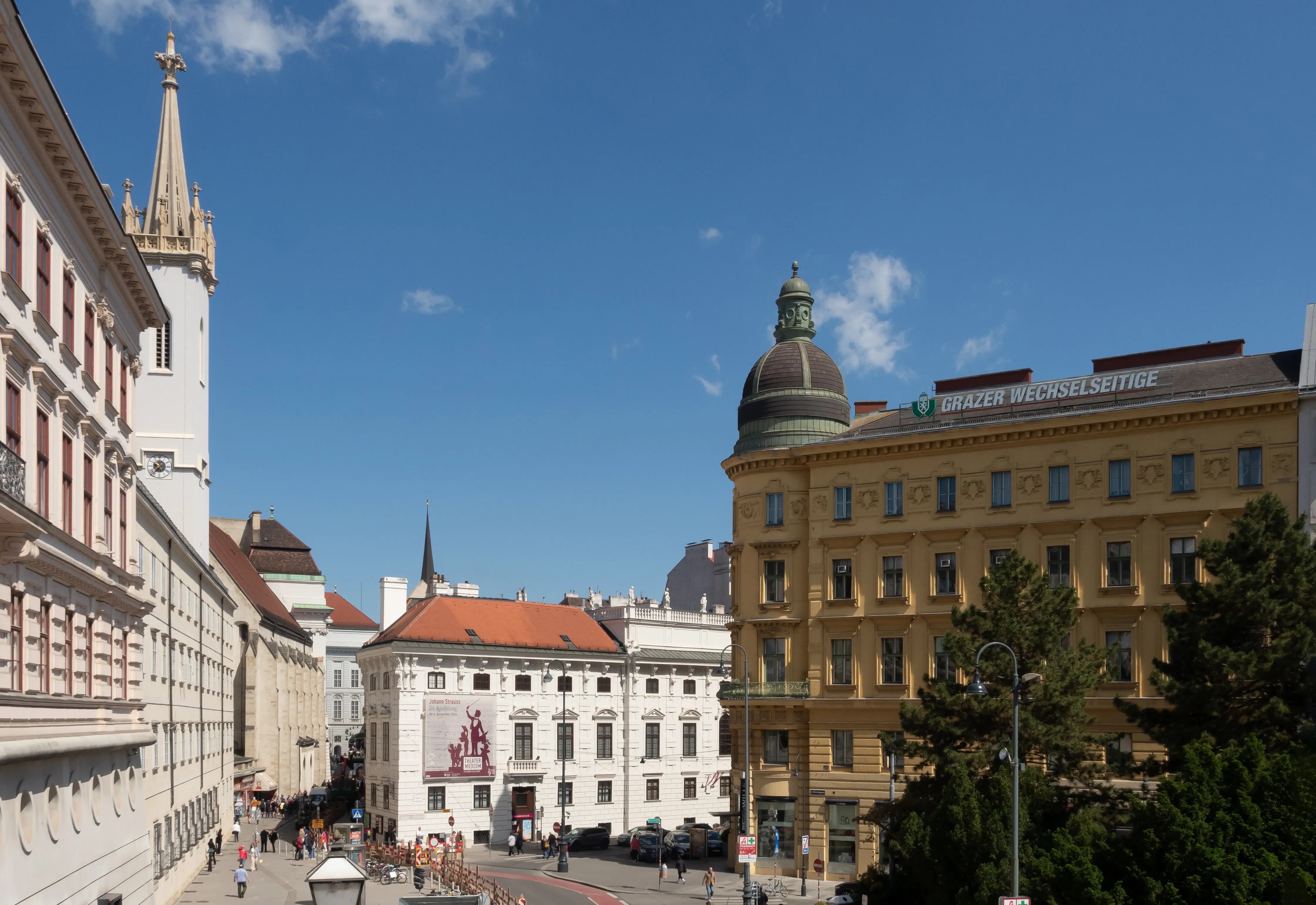 A tall white church spire with a golden cross stands next to a yellow building with a dome, under a blue sky with scattered clouds.