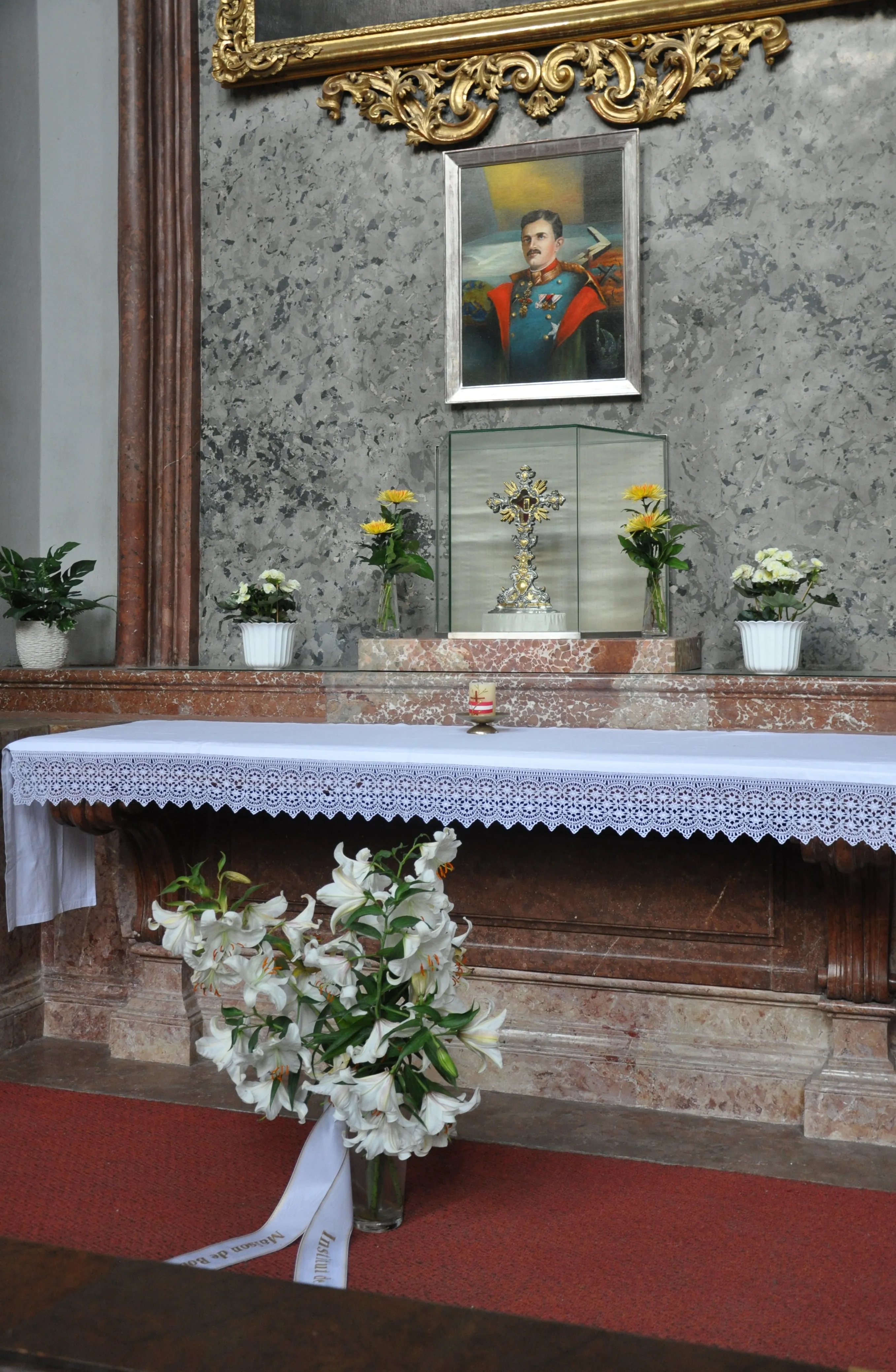 Altar with a portrait above, flowers, and a glass case containing a cross inside a church