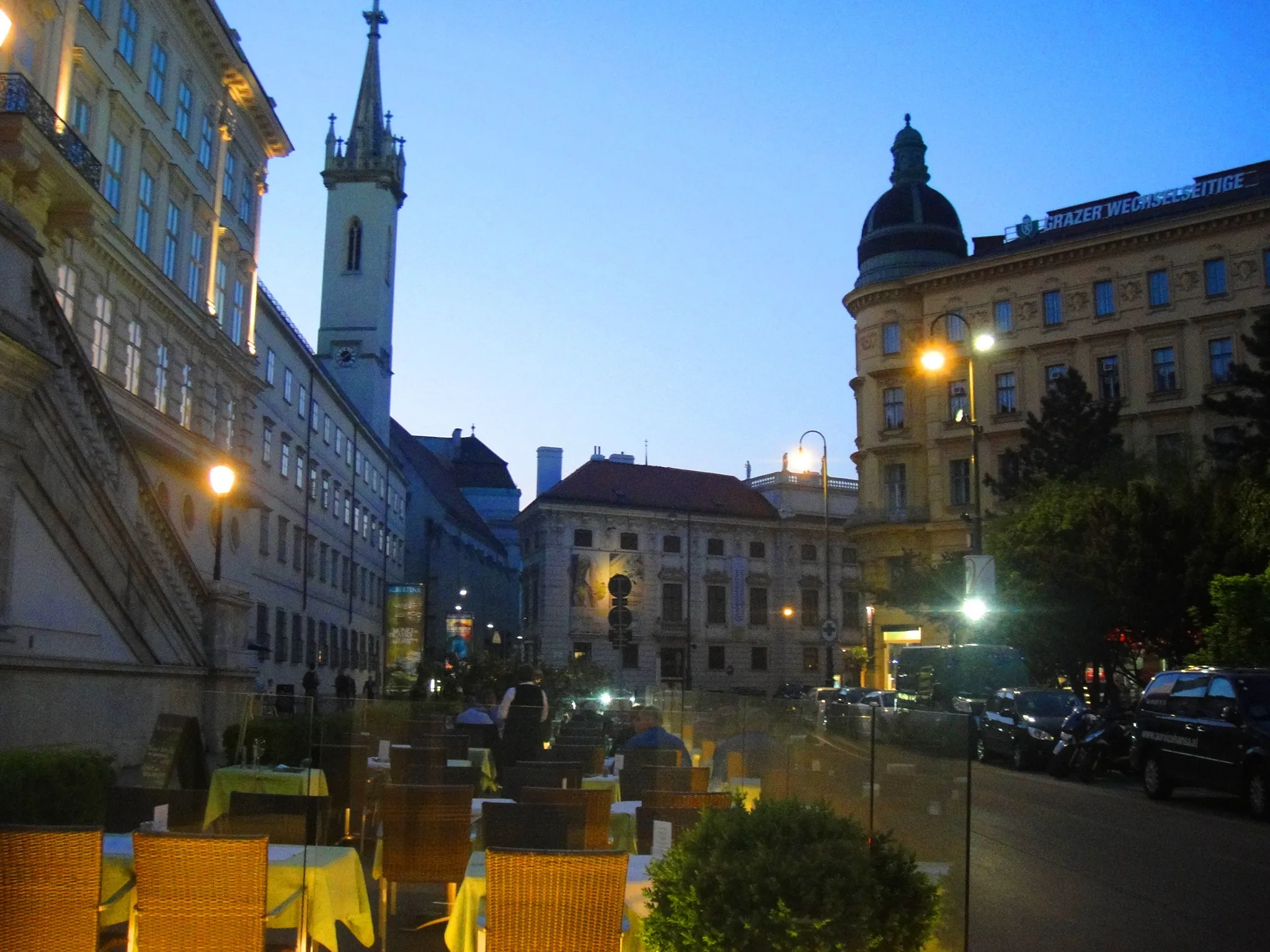Evening view of a historic street with a tall church spire, buildings with lit windows, and outdoor seating with tables and chairs.