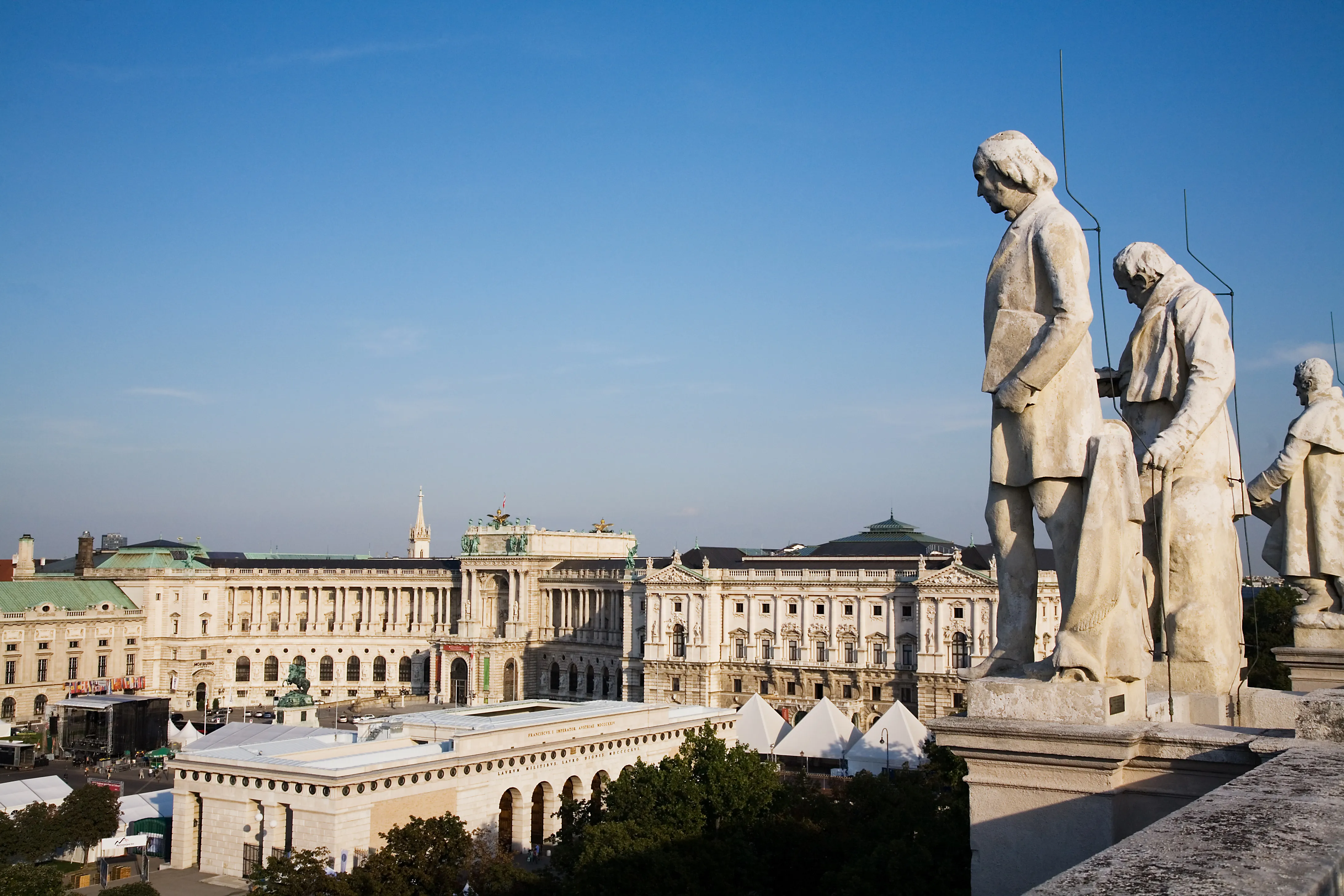 Statues of composers on a terrace with the Augustinian Church and Hofburg Palace in the background under a clear blue sky