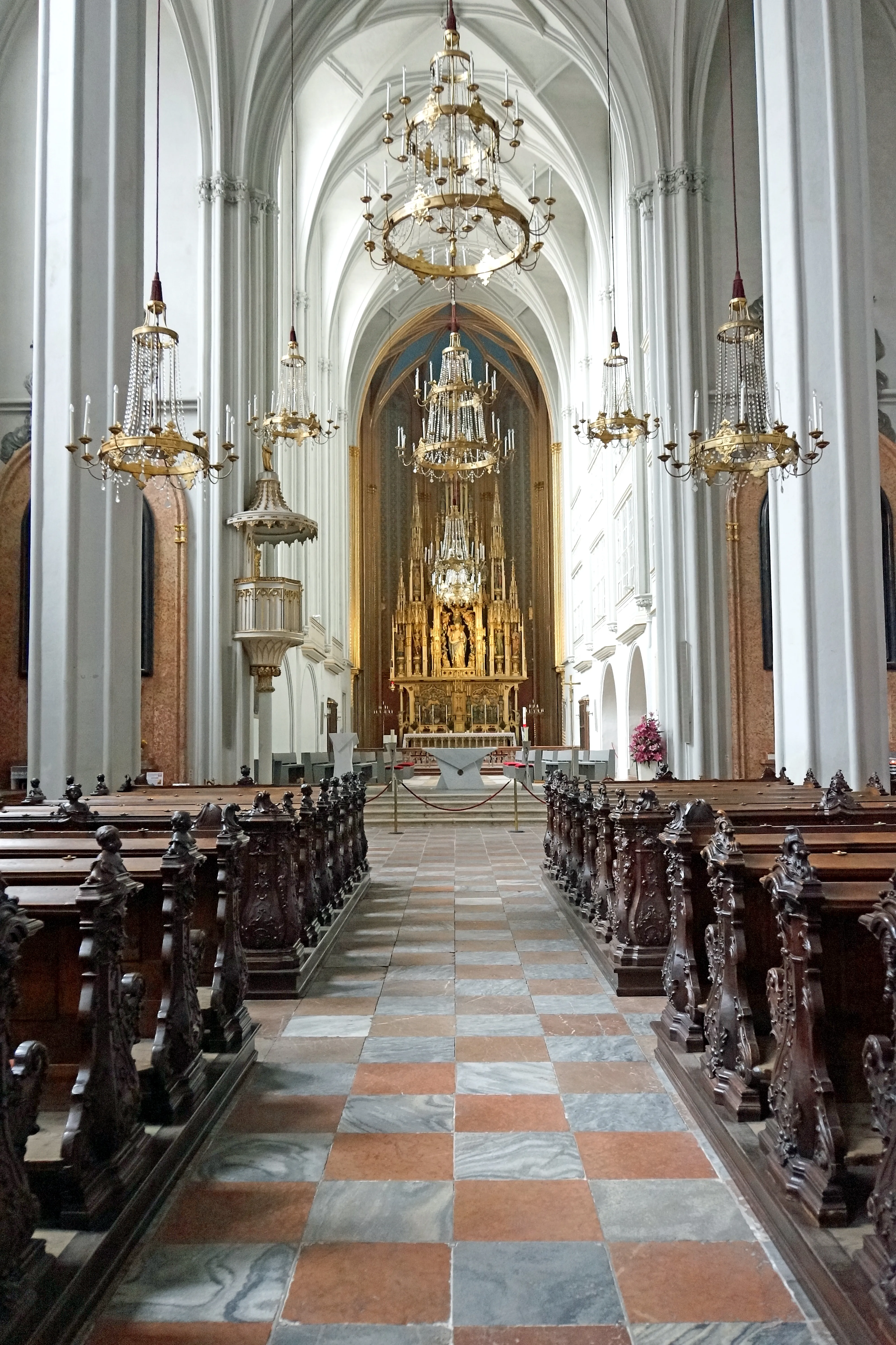 Interior of a church with high white columns, ornate chandeliers, wooden pews, and a checkered marble floor leading to a golden altar.
