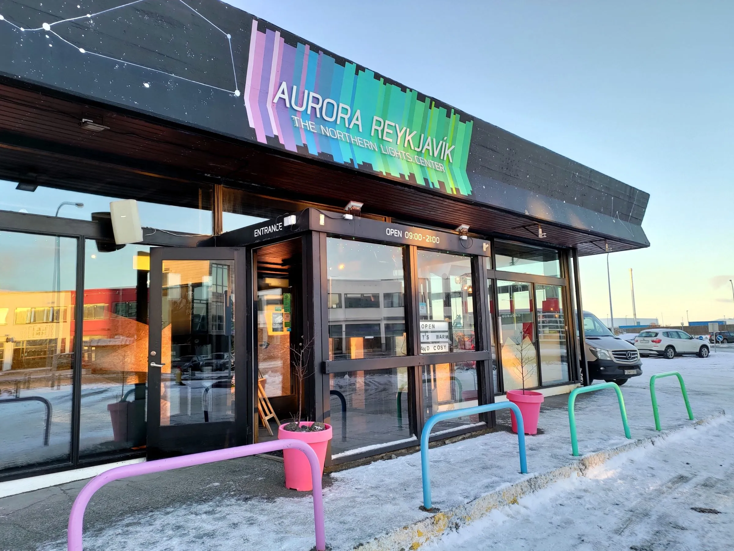 Widok z zewnątrz na wejście do Aurora Reykjavík The Northern Lights Center, z kolorowymi stojakami na rowery i śniegiem na ziemi.