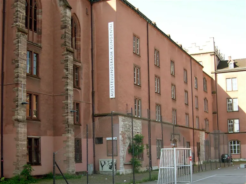 Buitenaanzicht van de Ausstellungsraum Klingental, een groot roodbruin gebouw met een verticale banner waarop de naam van de locatie staat, met een omheinde binnenplaats en een voetbaldoel aan de voorzijde.