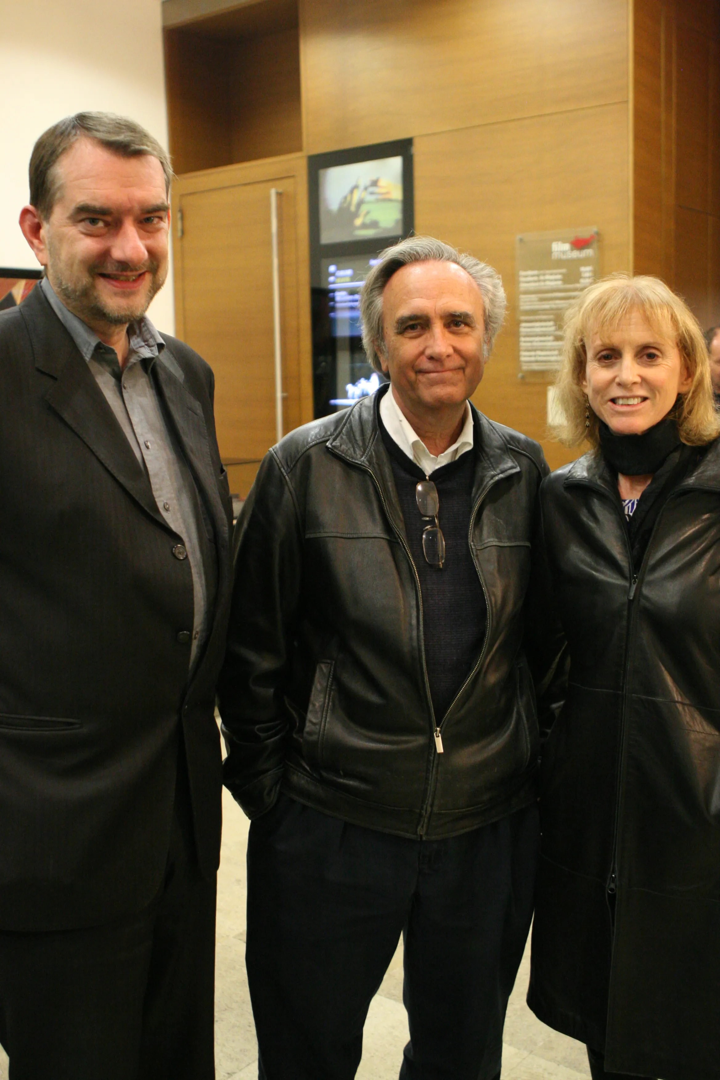 Three people standing together inside a building with wooden walls and a screen displaying a film scene.