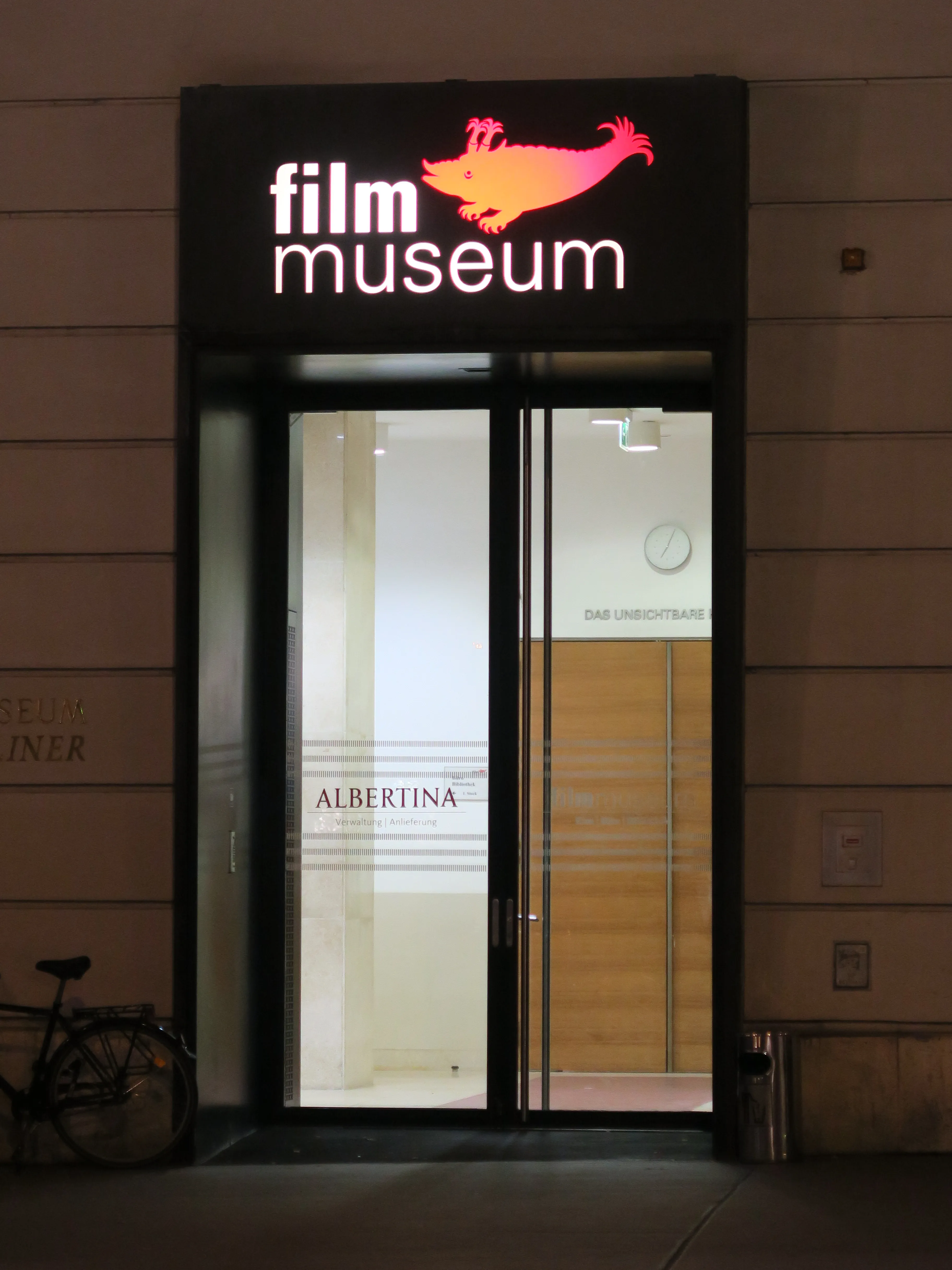 Illuminated sign above glass doors reading 'film museum' with an orange bird logo, entrance to the Austrian Film Museum at night
