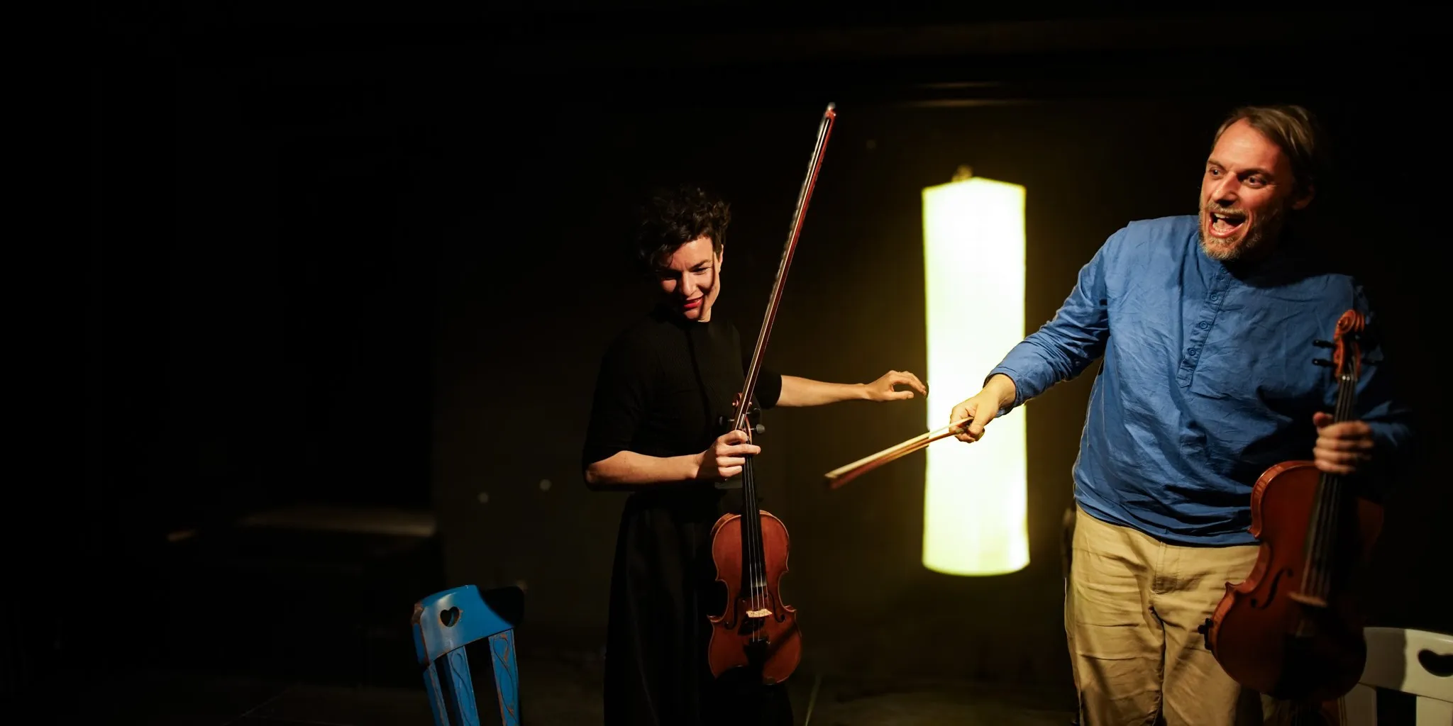 Dos músicos actuando con violines en un escenario con poca luz