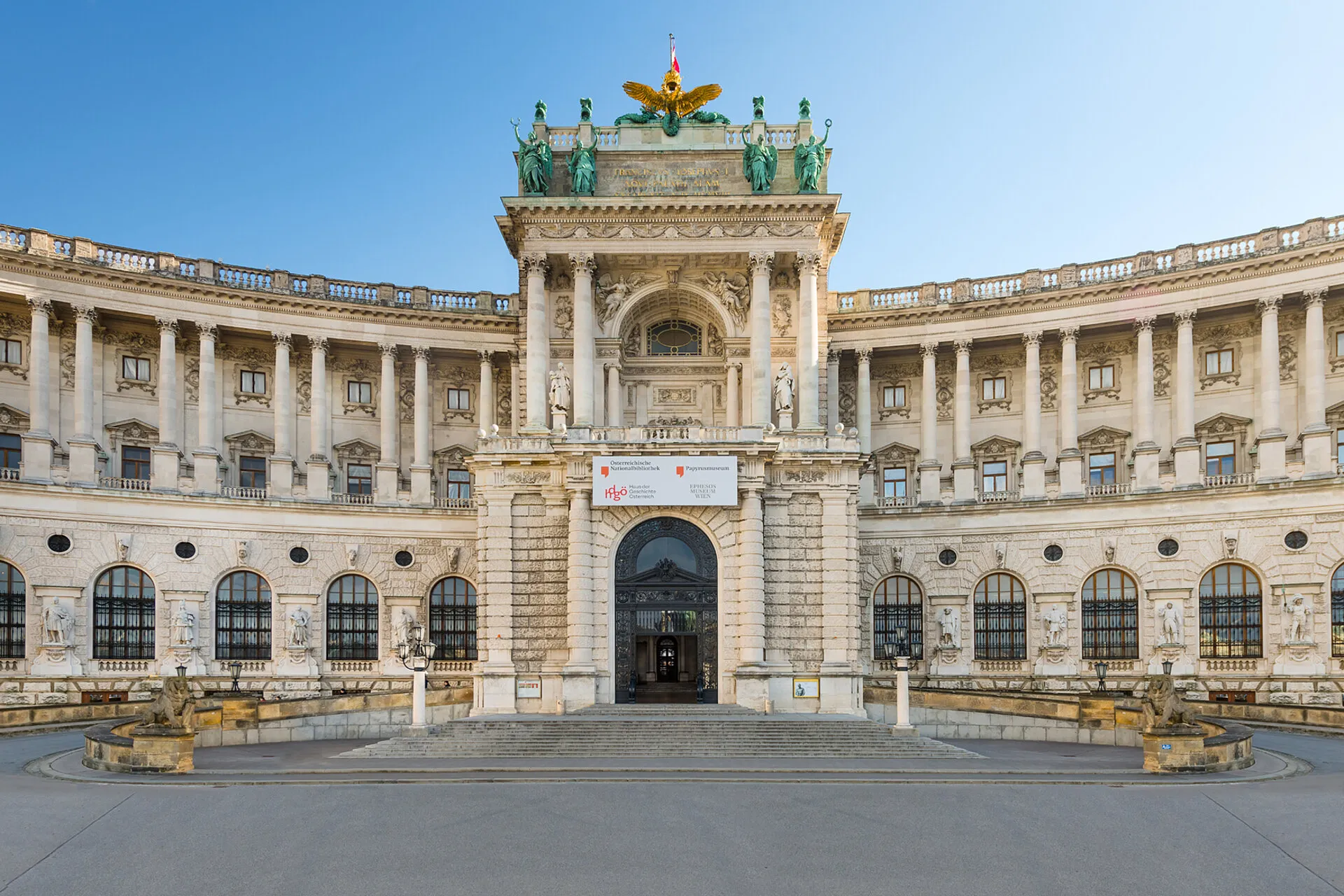 Fachada neoclásica de la Biblioteca Nacional de Austria con entrada central, ventanas arqueadas y un grupo escultórico en el tejado contra un cielo azul claro