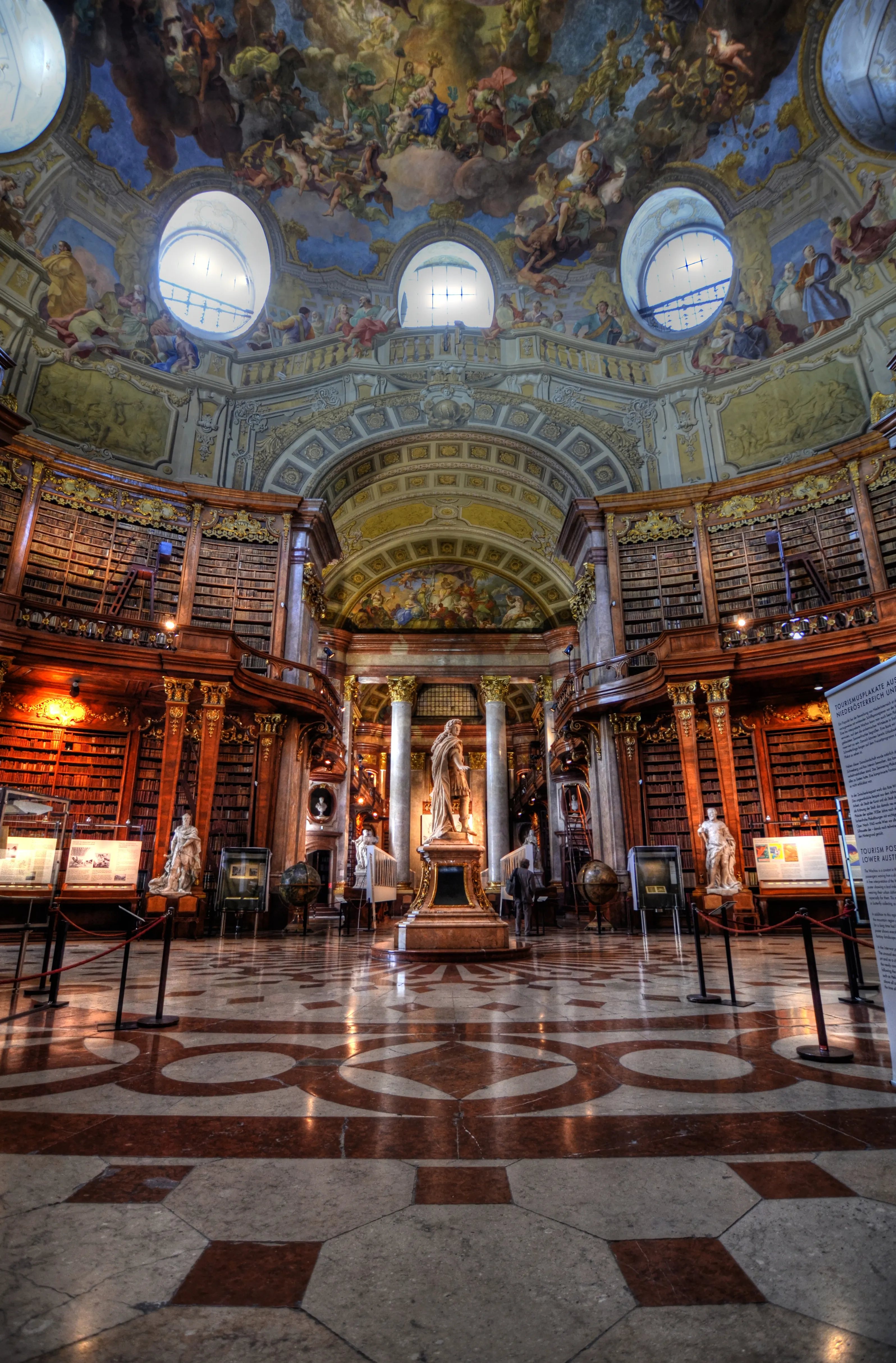 Interior del Salón de Estado de la Biblioteca Nacional Austriaca con un techo abovedado pintado con frescos, estanterías de madera, una estatua central y un suelo de mármol estampado