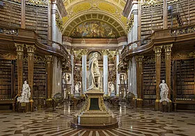 Gran interior de una biblioteca con altos techos abovedados, decoraciones doradas y hileras de estanterías flanqueando una estatua central.