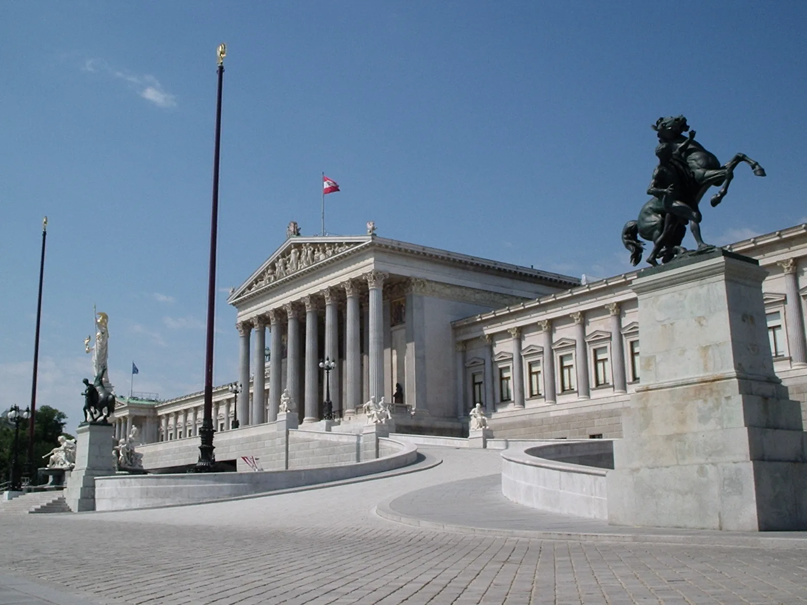 Edificio neoclásico con columnas y una bandera, flanqueado por estatuas sobre pedestales
