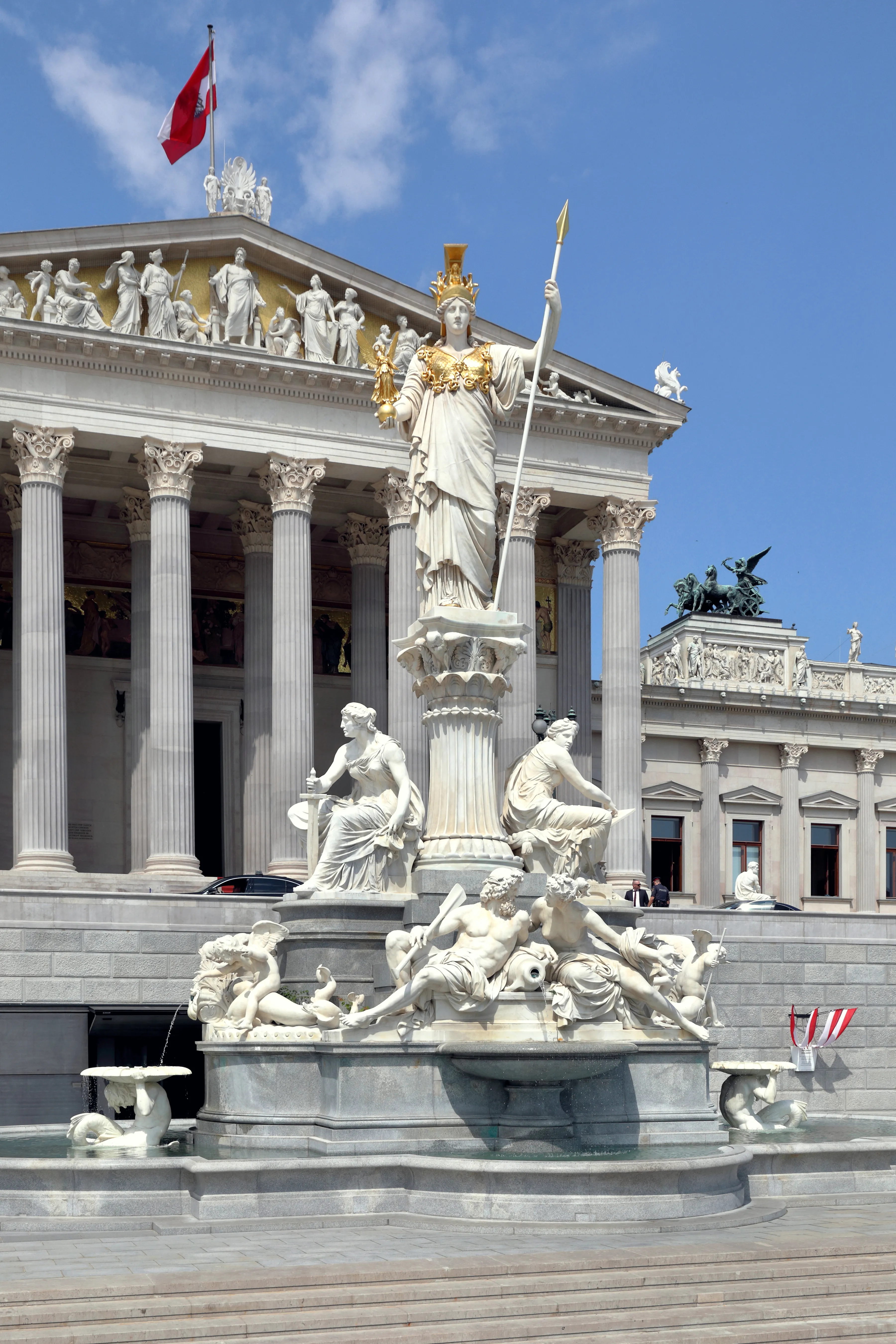 Neoclassicistisch gebouw met zuilen en een groot standbeeld van Pallas Athena op een fontein ervoor, onder een blauwe lucht met een wapperende rood-witte vlag