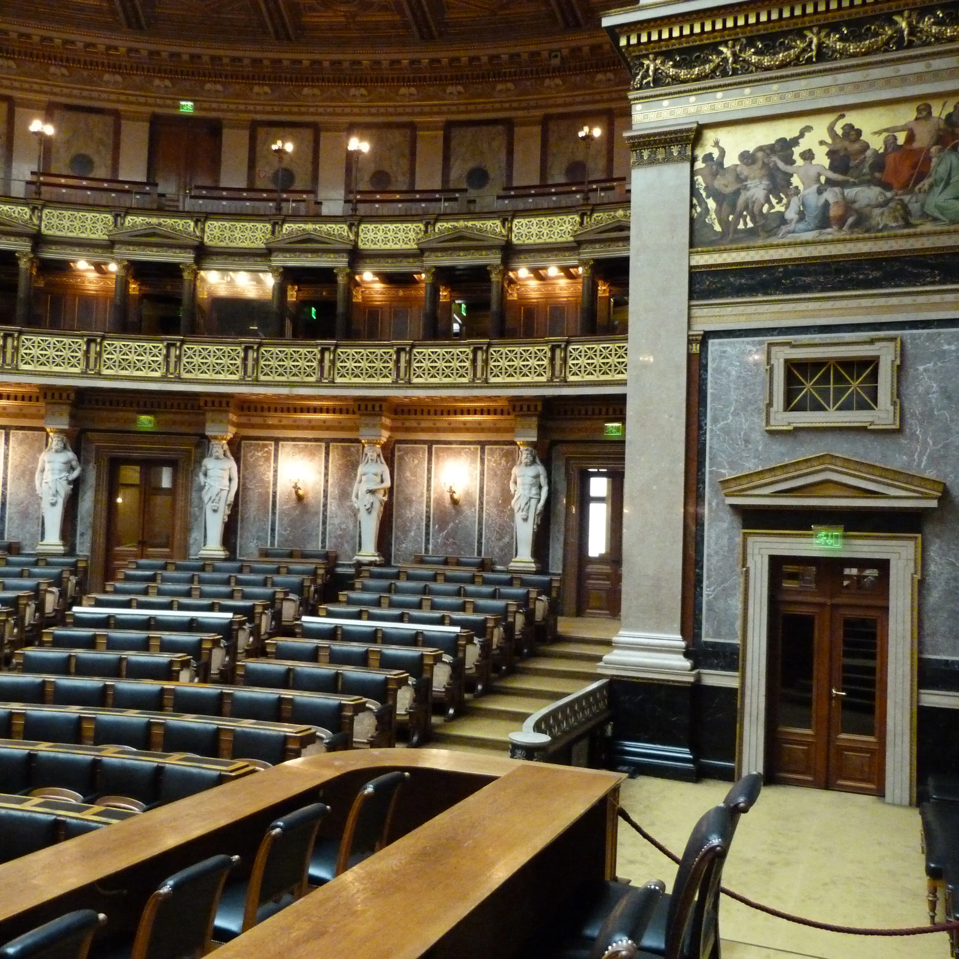 Interieur van een grote zaal met rijen stoelen, sierlijke balkons, beelden en een muurschildering aan de muur