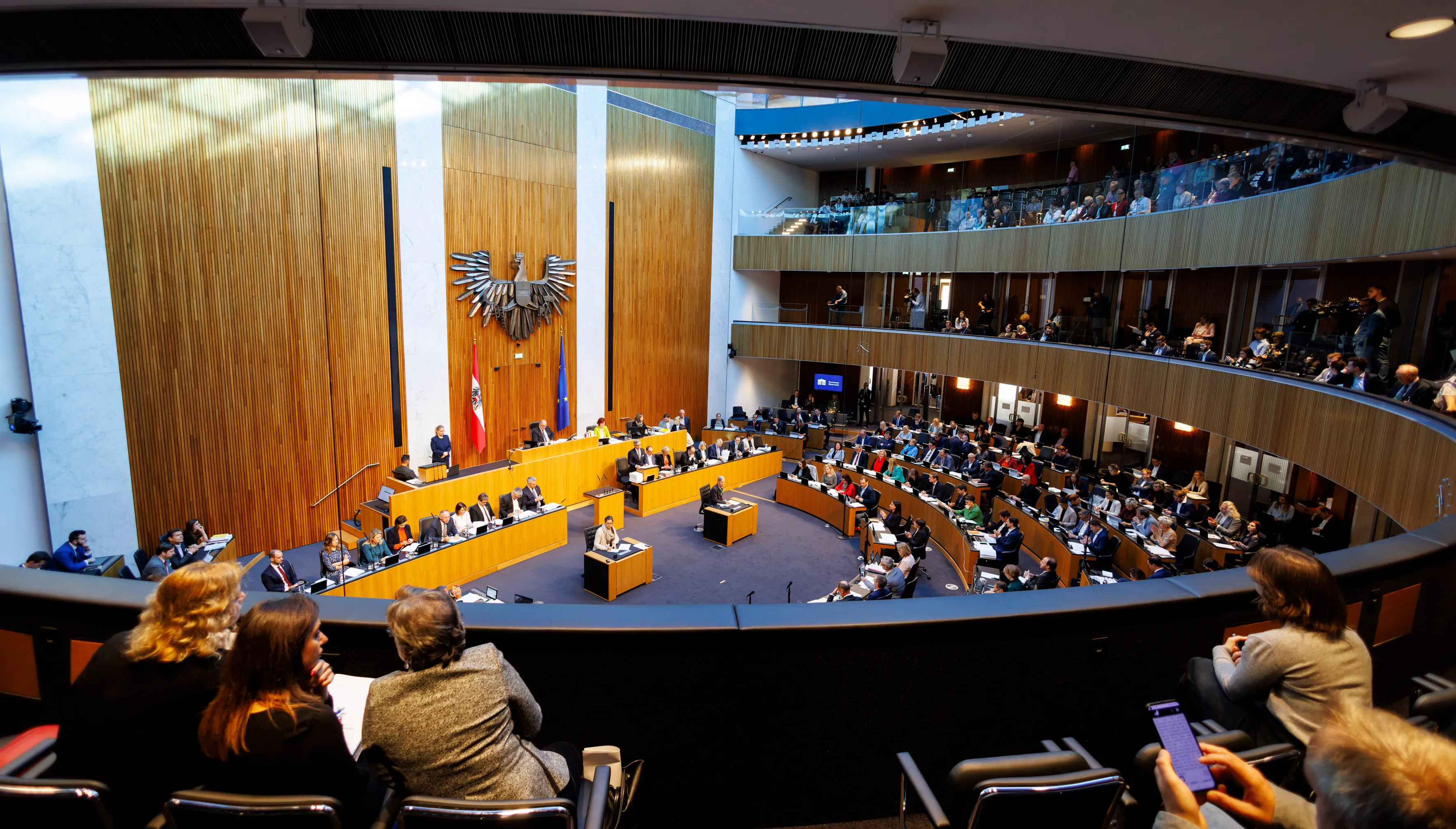 Interieur van de Oostenrijkse parlementszaal met rijen bureaus, zittende mensen en een groot adelaarembleem op de muur