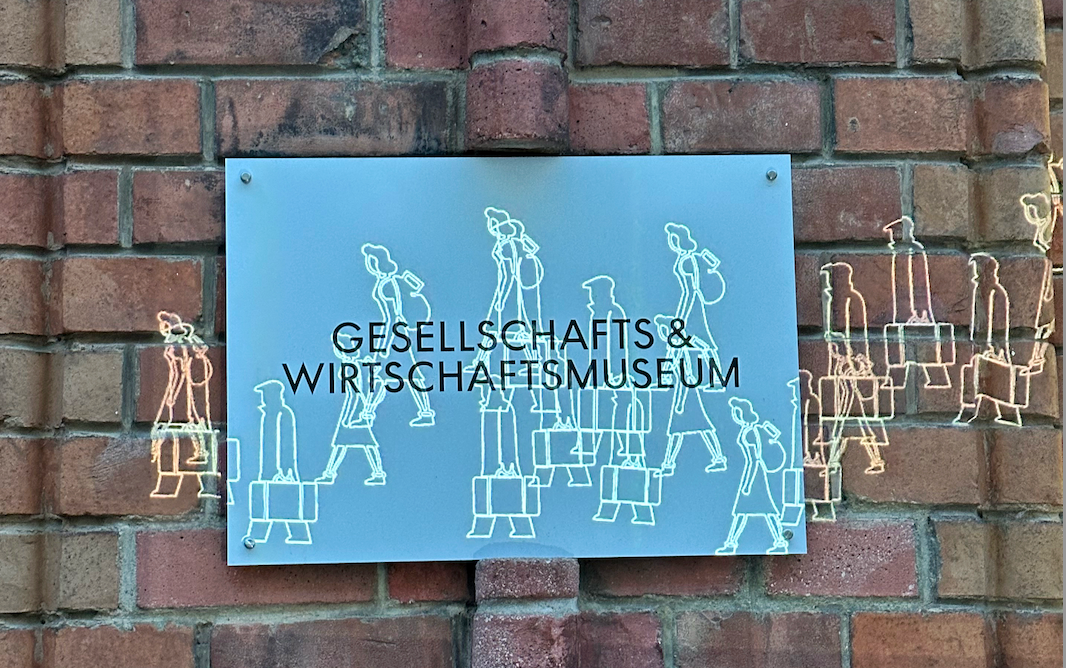 Blue sign with white text 'GESELLSCHAFTS & WIRTSCHAFTSMUSEUM' and silhouettes of people on a brick wall