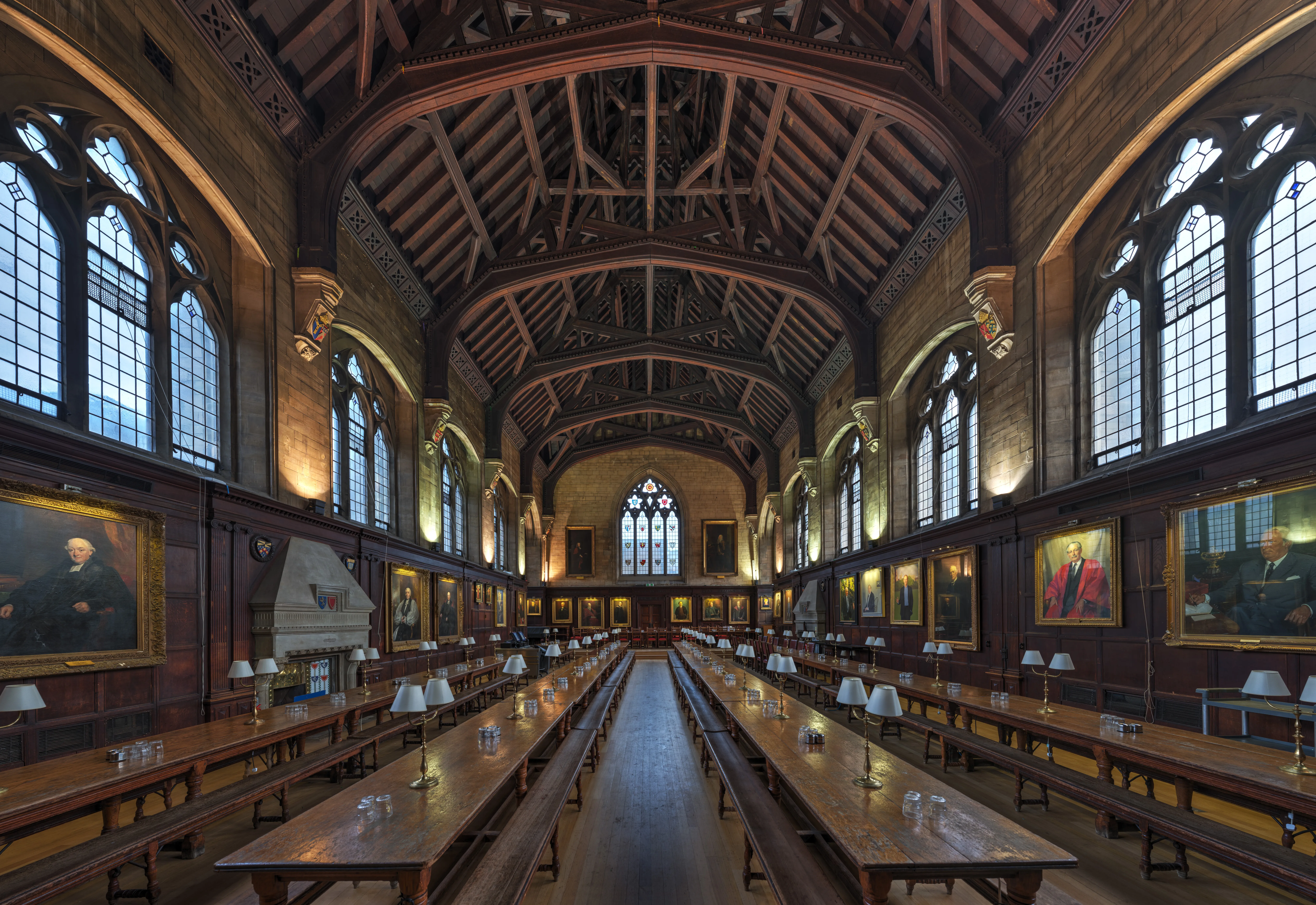 Largas mesas de comedor de madera se extienden por el centro del gran salón de Balliol College, flanqueadas por retratos y ventanas góticas arqueadas bajo un techo de madera abovedado.