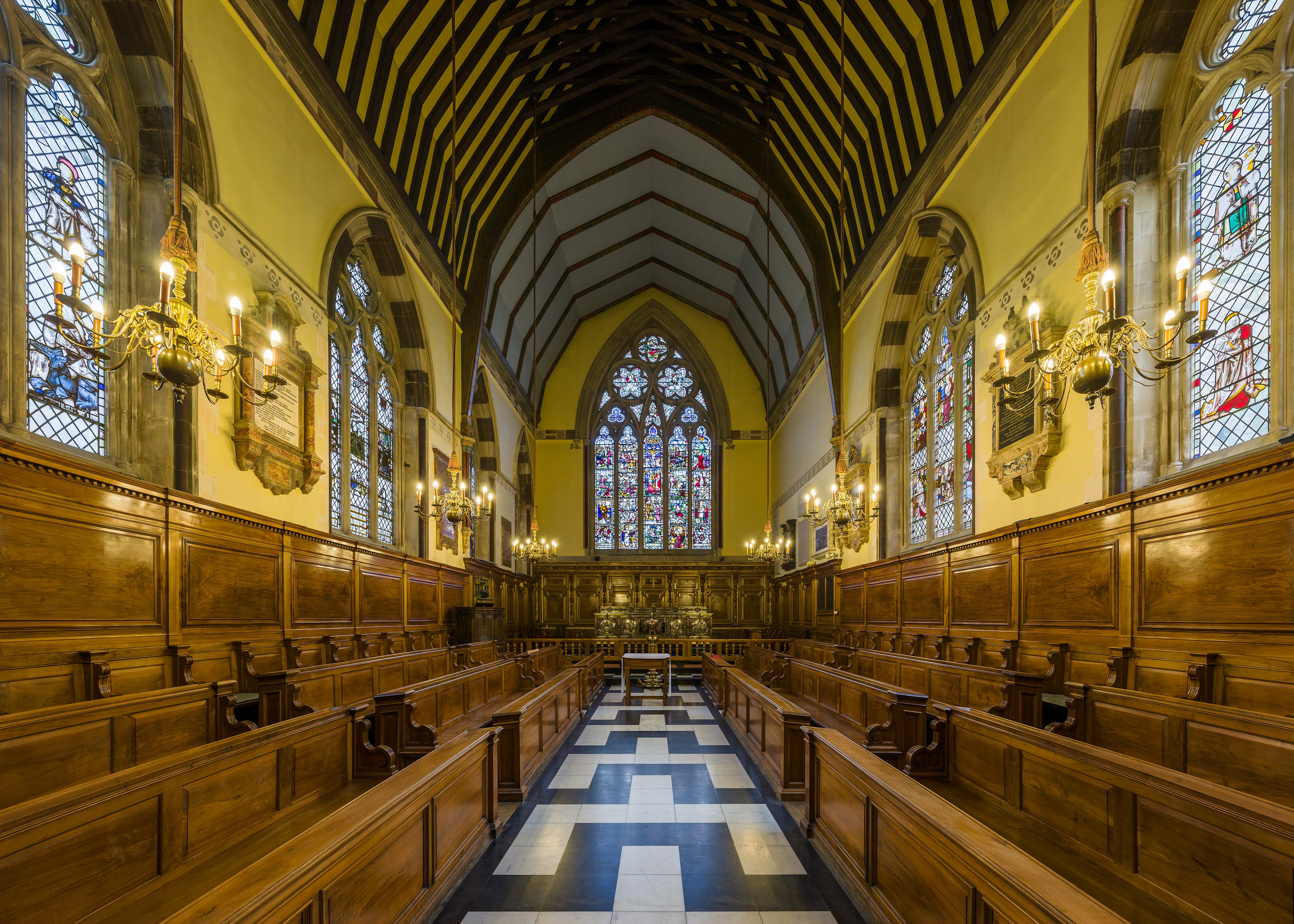 Interior de la capilla del Balliol College que muestra filas de bancos de madera, un suelo de damero en blanco y negro, vidrieras, candelabros montados en la pared y un techo abovedado con vigas de madera oscura.