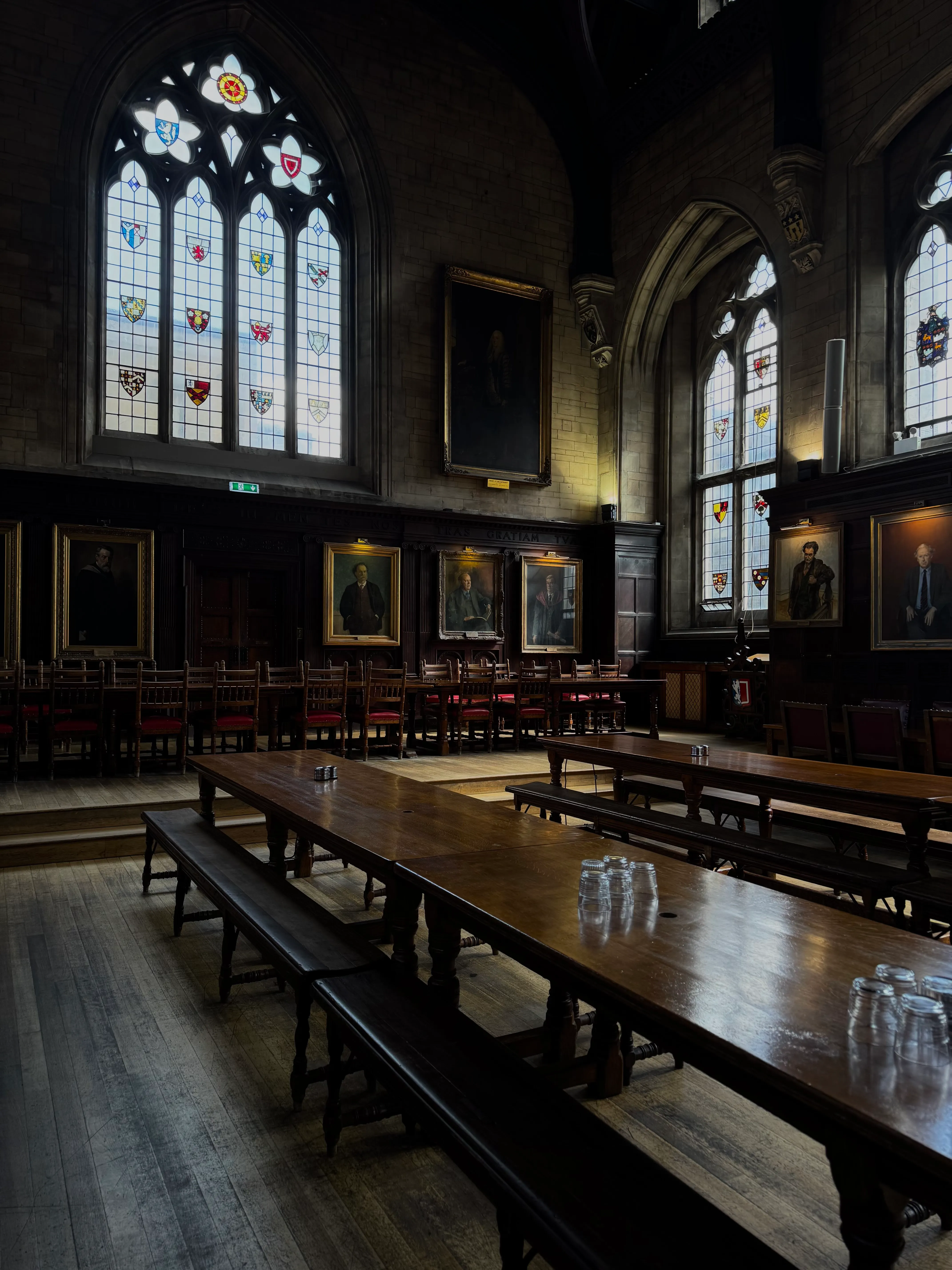 Interior del comedor de Balliol College, que presenta largas mesas y bancos de madera, grandes vidrieras con escudos heráldicos y retratos enmarcados en paredes revestidas de madera oscura.