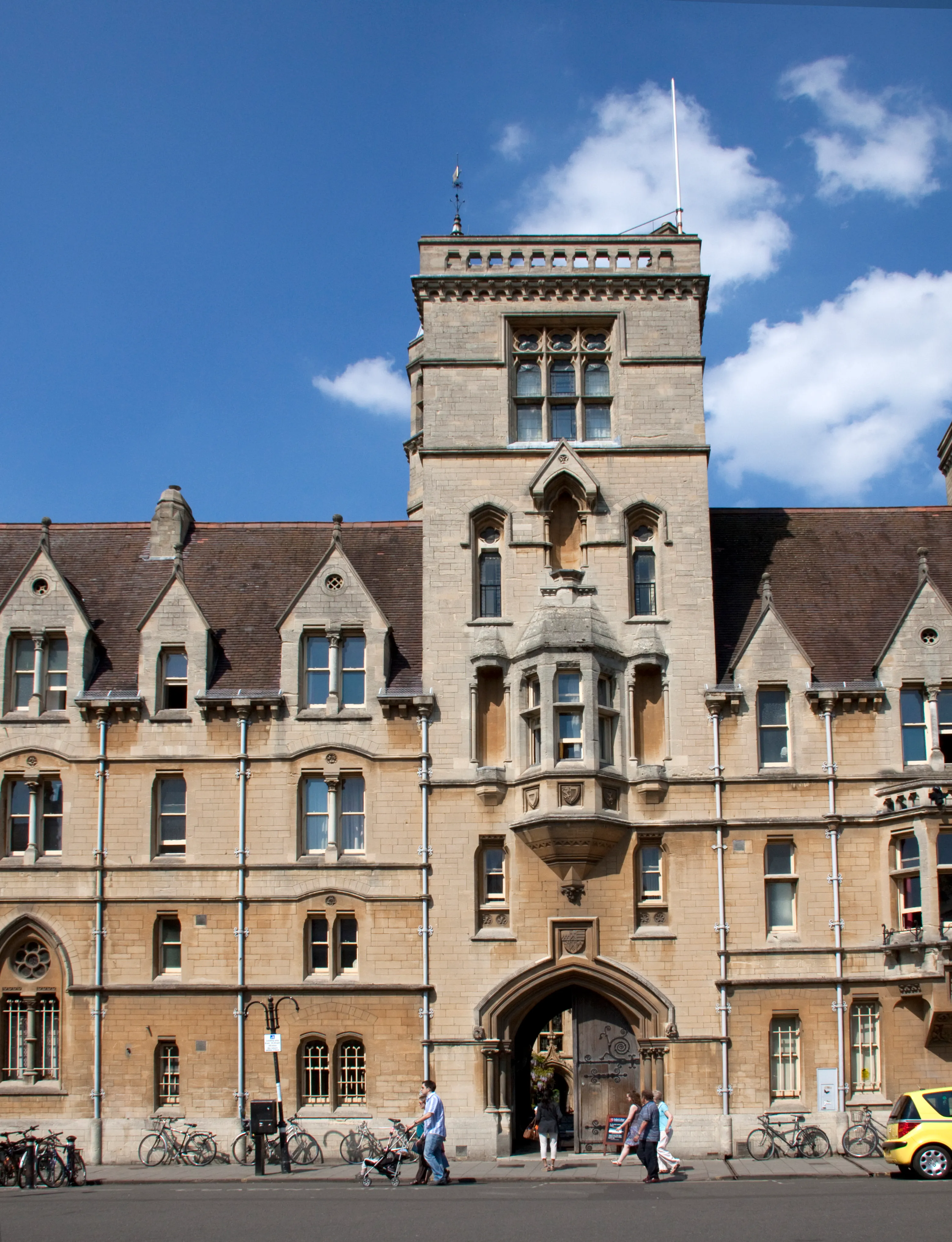 Fachada de piedra del Balliol College de Oxford que presenta una torre central con entrada arqueada, ventanas góticas y bicicletas aparcadas a lo largo de la calle.