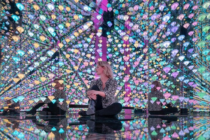 Luces coloridas en forma de diamante suspendidas en una sala espejada con visitantes sentados en el suelo